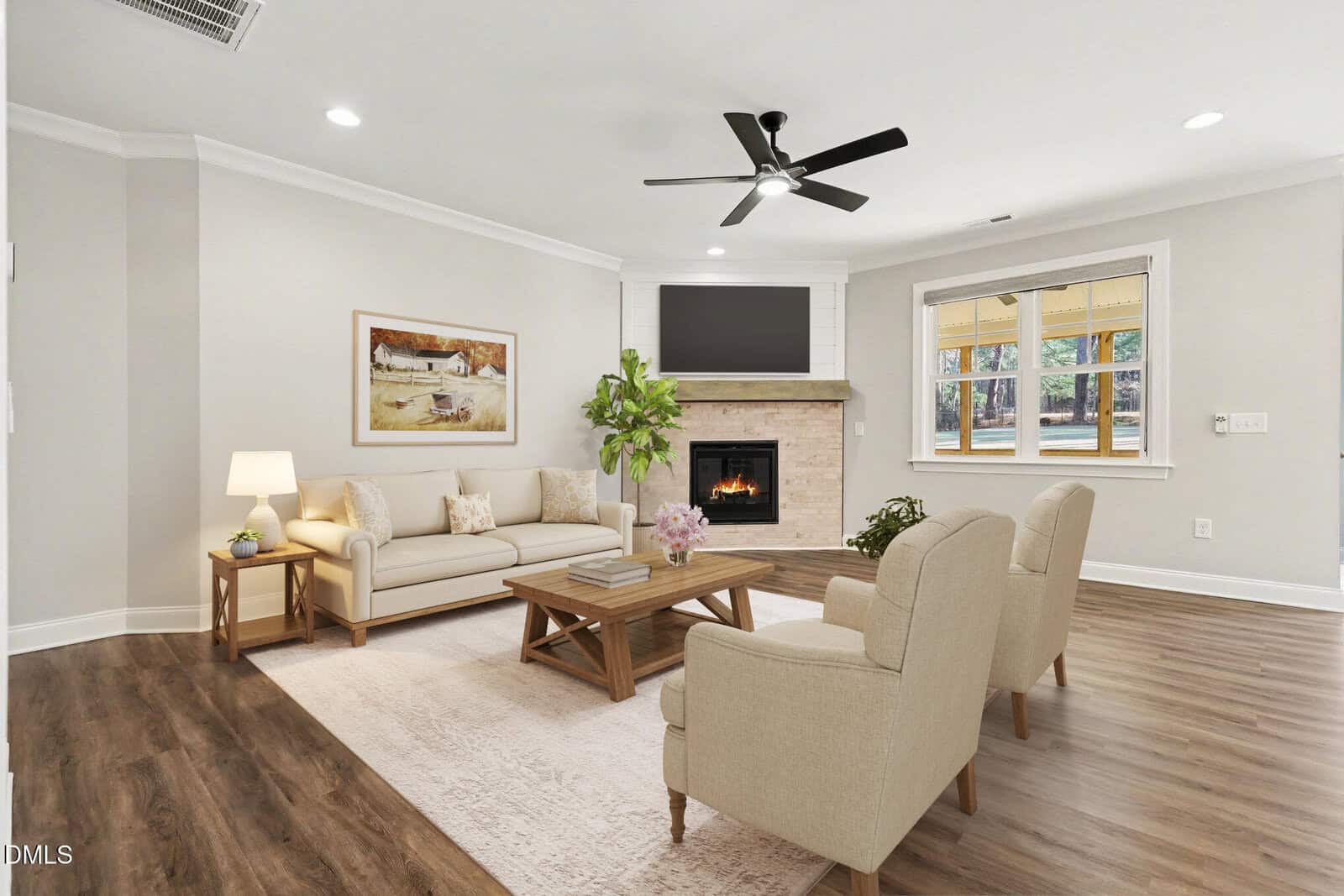 A bright living room with wood floors, a beige sofa, two matching armchairs, a wooden coffee table, a fireplace with a TV above, a side table with a lamp, a framed painting, a potted plant, and large windows.