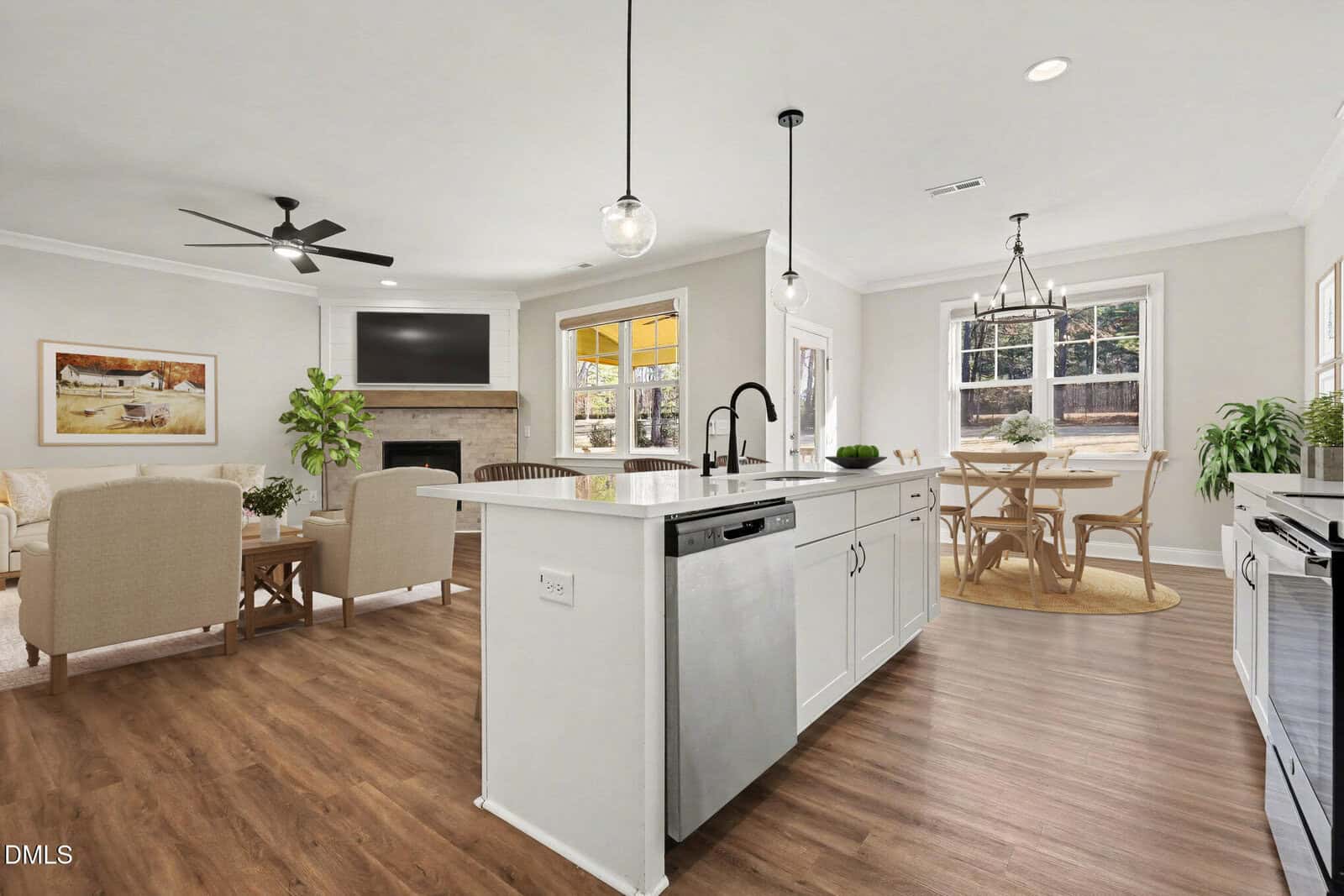Open concept kitchen and living area with white cabinets, island with sink, dining table by window, cozy seating, wood floors, ceiling fan, and light neutral decor. Large windows let in natural light.