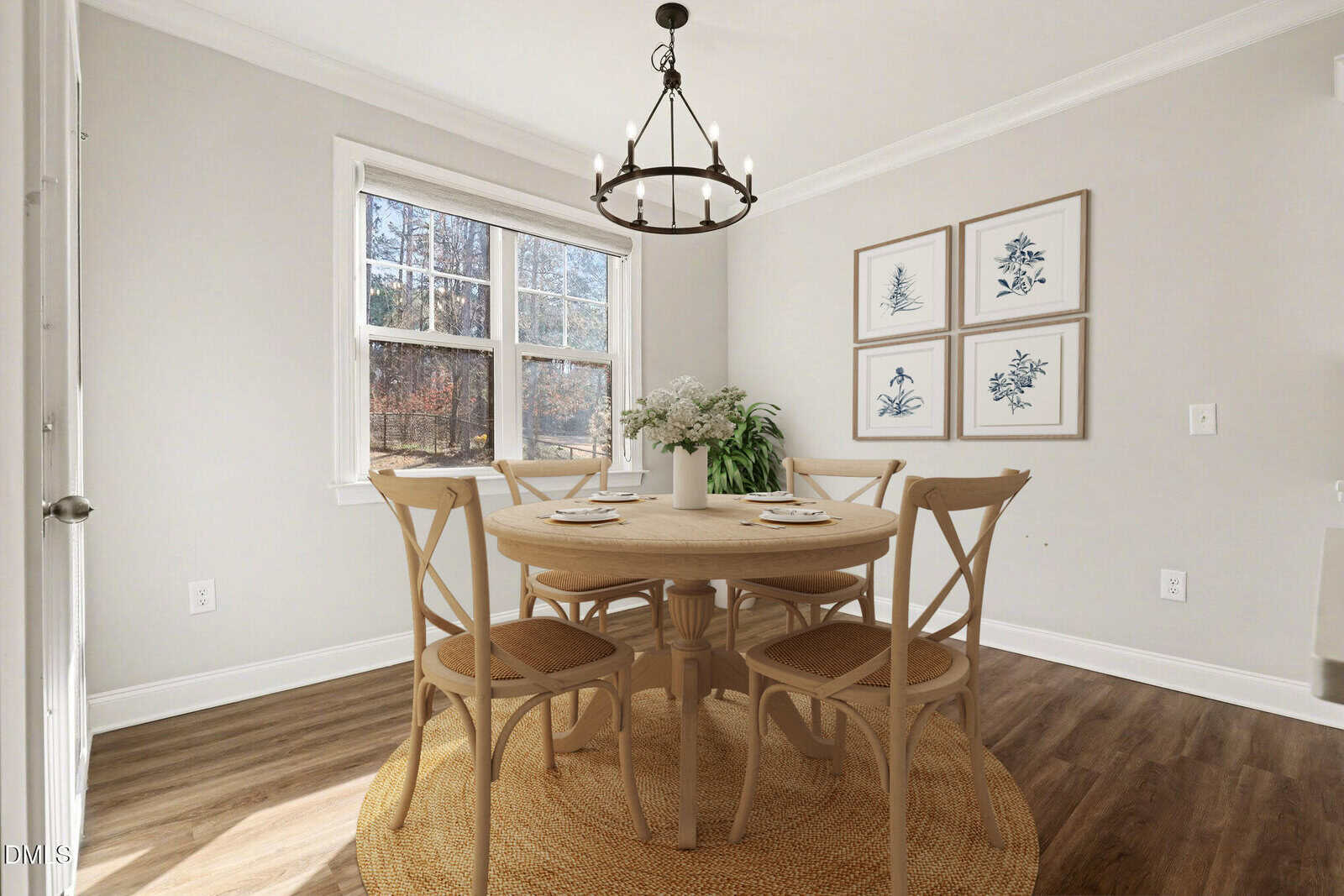 A bright dining room with a round wooden table, four chairs, a woven rug, a floral centerpiece, and botanical prints on the wall. A window lets in natural light, highlighting the wood floor and neutral walls.