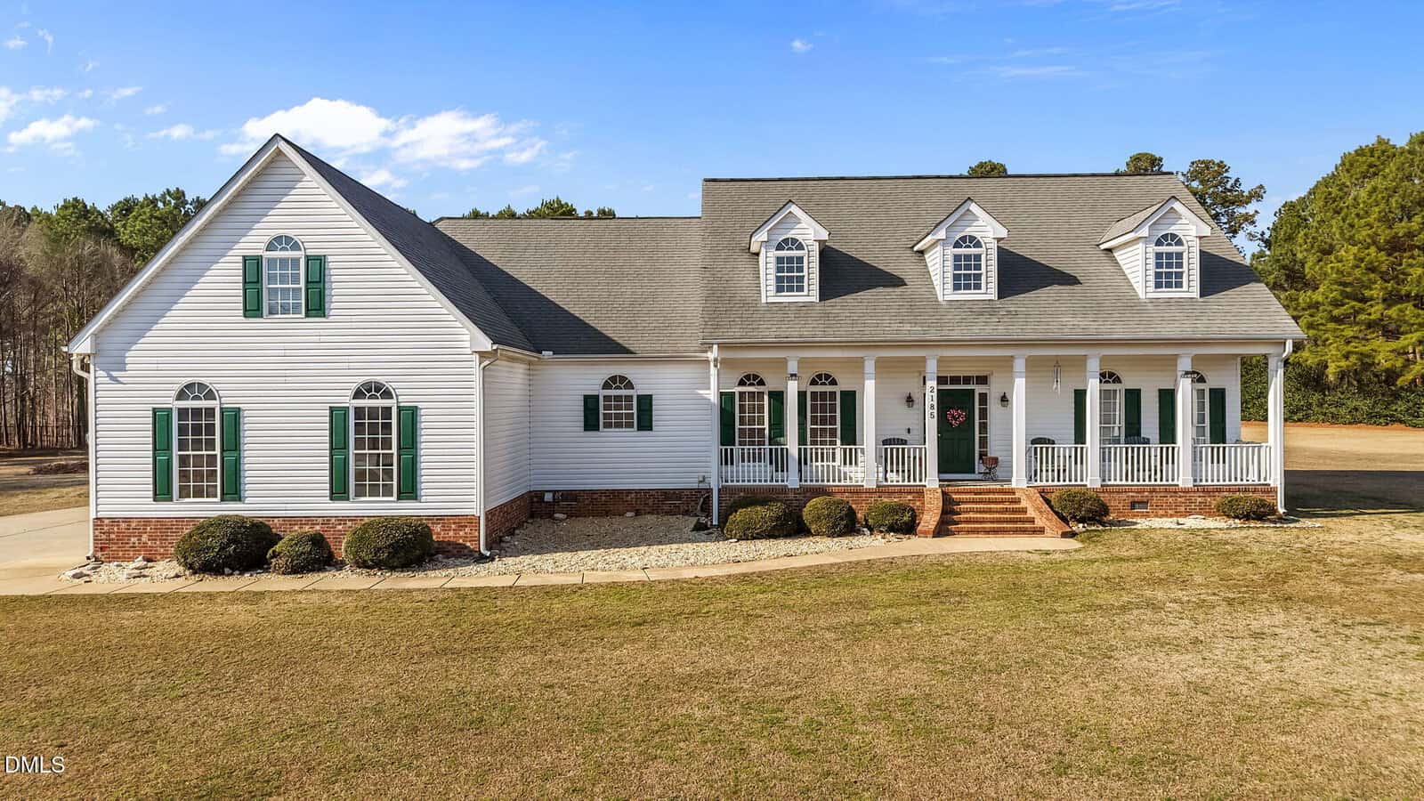A large white two-story house with green shutters, brick foundation, and a covered front porch with columns. The house has three dormer windows on the roof and sits on a spacious lot with minimal landscaping.