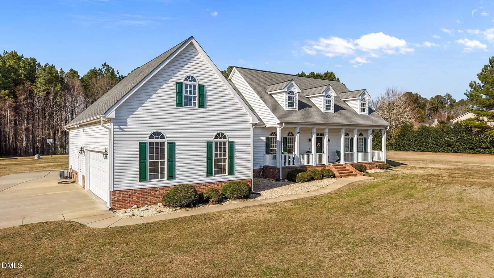 A large white house with green shutters, brick accents, and a covered front porch sits on a spacious lawn with minimal landscaping and a driveway, bordered by trees under a blue sky.