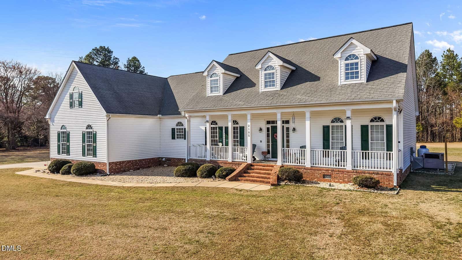 A white, two-story house with green shutters, a gray roof, dormer windows, and a large front porch with columns and railings. A small brick walkway leads up to the entrance. The lawn is spacious and mostly bare.