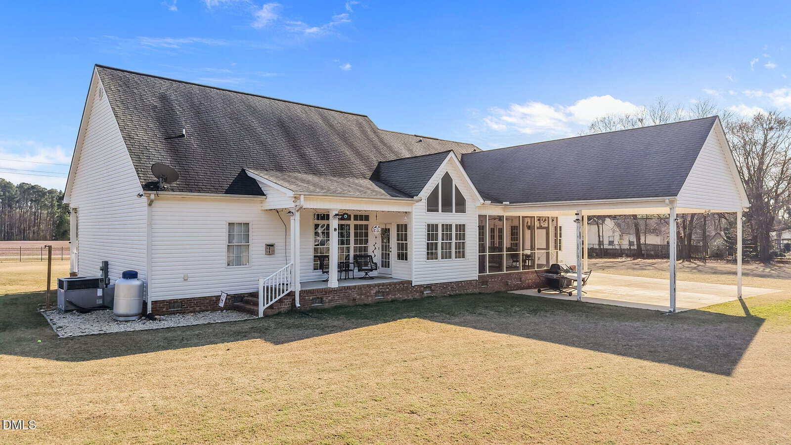 A white single-story house with a covered porch and carport, large windows, and a shingled roof sits on a grassy lawn under a clear blue sky. Outdoor seating and a grill are visible on the porch.
