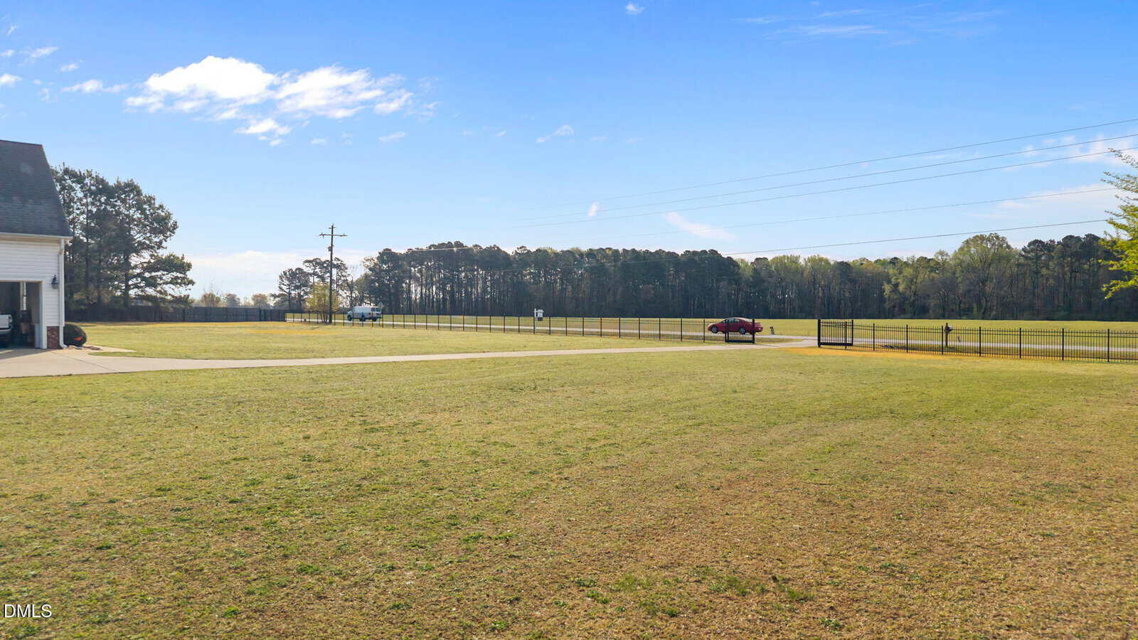 A wide, open grassy yard with a driveway on the left, a few cars parked near a fence, and trees lining the background under a mostly clear sky.