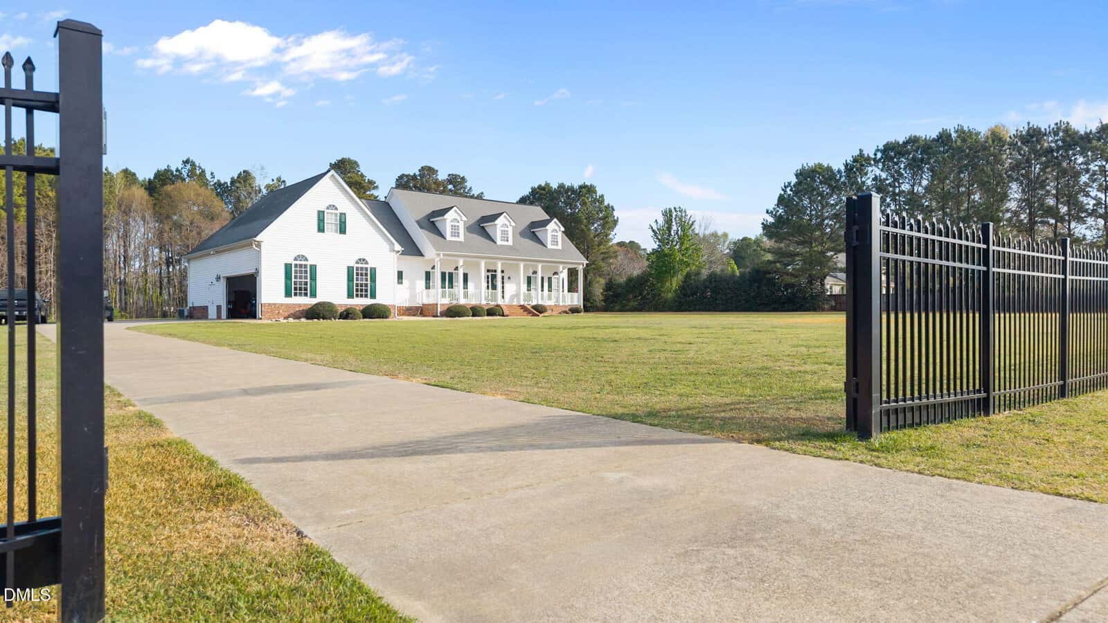 A large white house with dormer windows and a front porch sits behind a black iron fence on a spacious lawn with a long concrete driveway, bordered by trees under a clear blue sky.