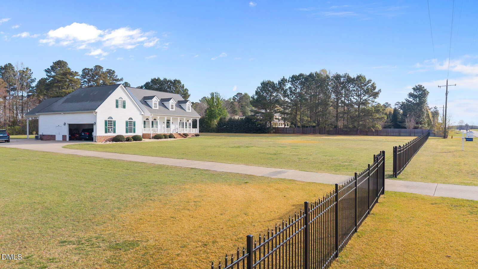 A large white house with black shutters sits on a spacious grassy lot with a long driveway and a black metal fence in the foreground. Tall trees line the back of the property under a clear blue sky.