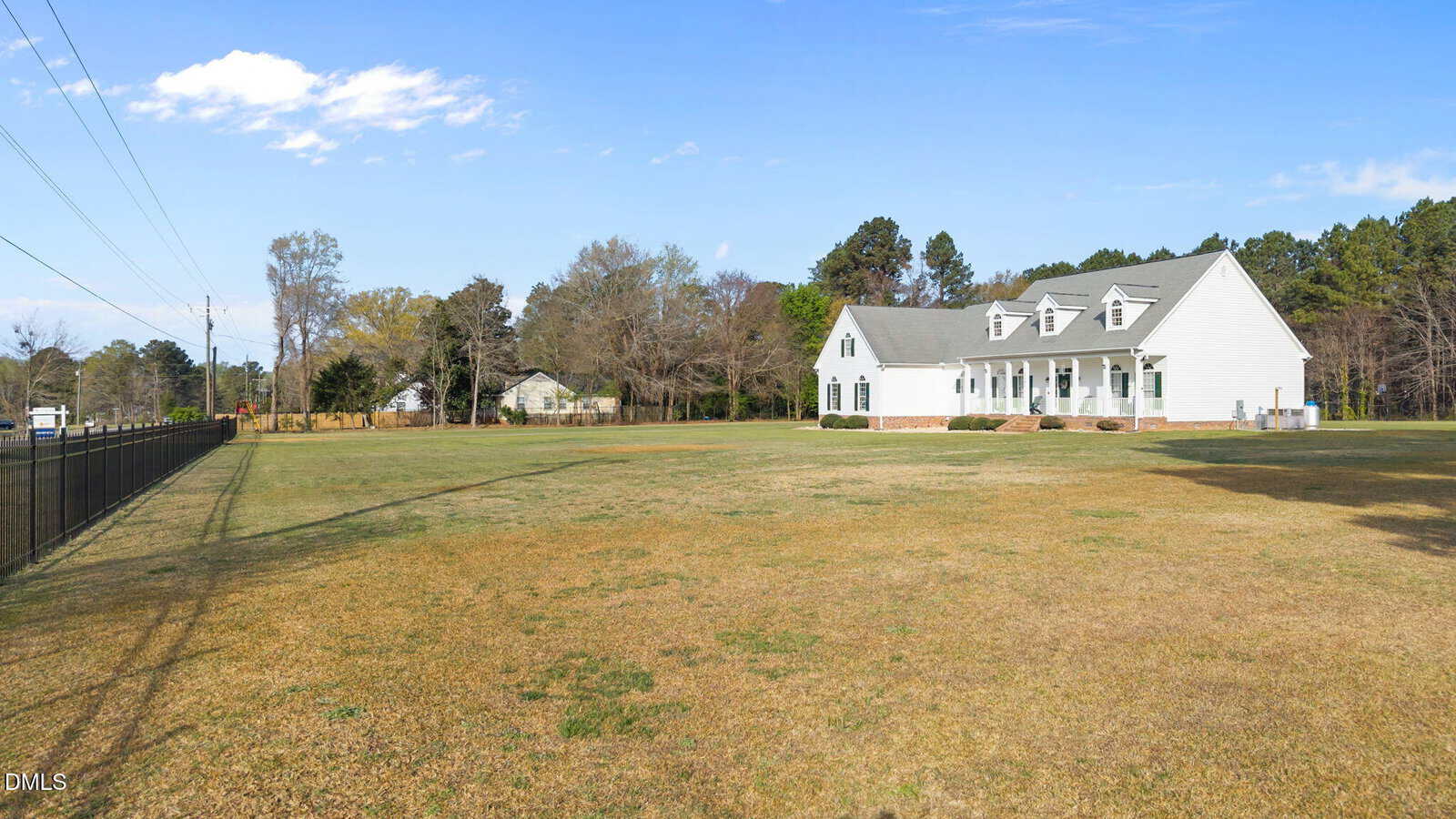 A large, white house with multiple windows and dormers sits on a spacious, grassy yard bordered by a black metal fence, with trees and another house visible in the background under a clear sky.
