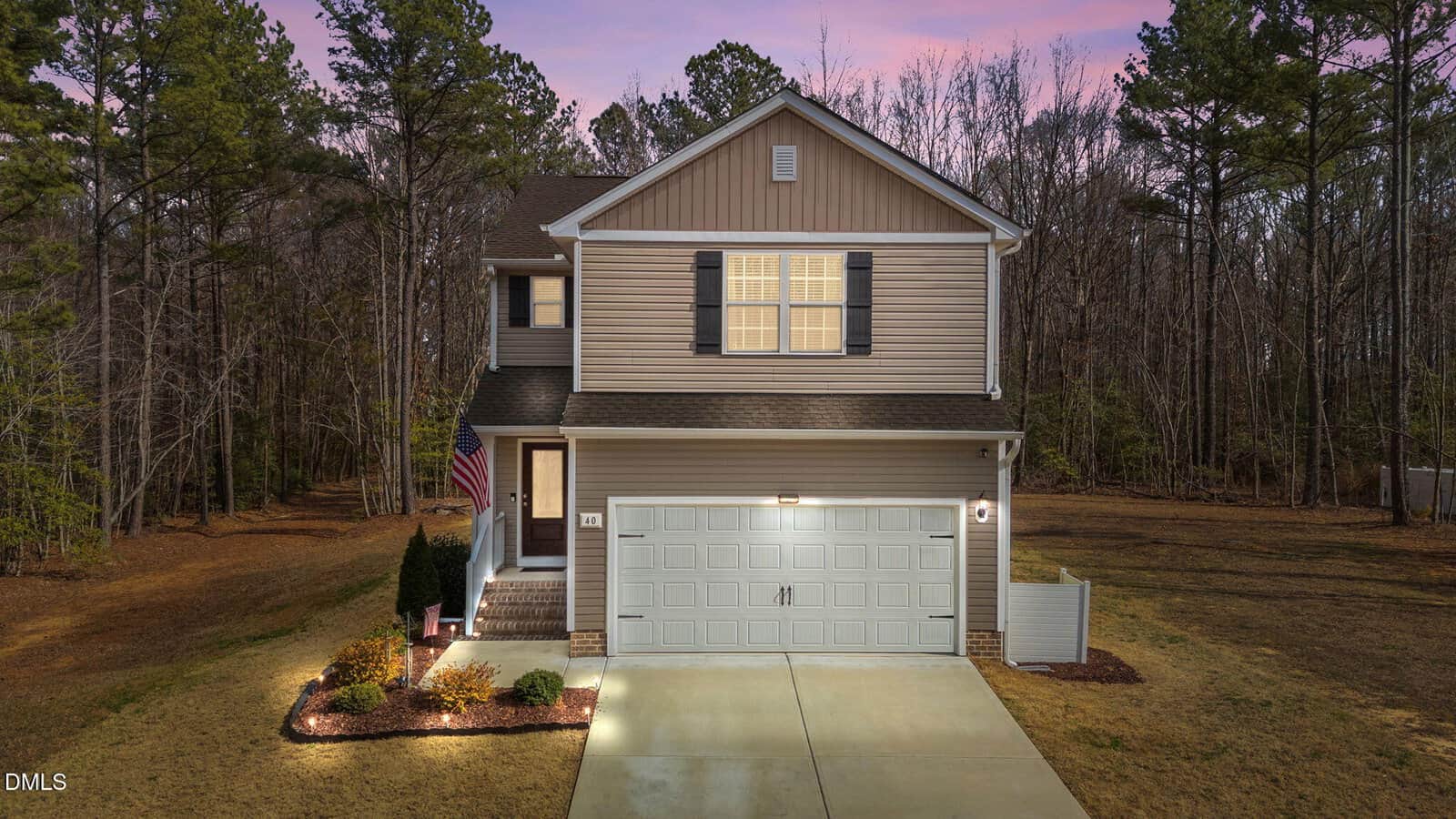 Two-story suburban house with beige siding, white trim, and a double garage. Lights are on inside and along the walkway. An American flag hangs near the front door. The house is surrounded by trees and lawn at dusk.