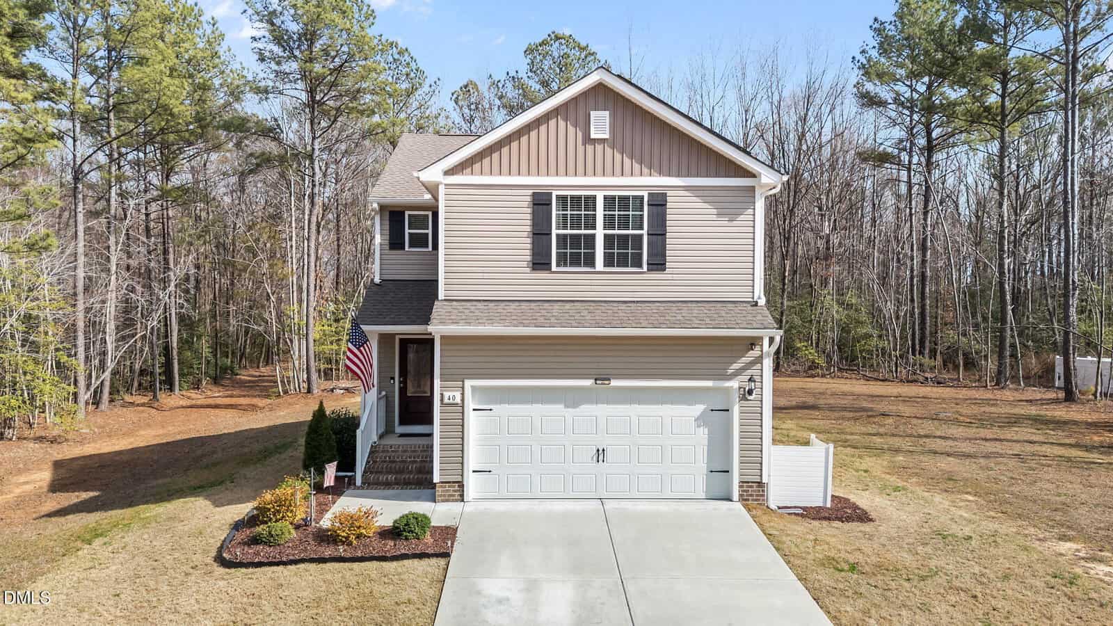 A two-story beige house with white trim, a double garage, and black shutters. The house has a front porch with steps, an American flag, and is surrounded by a lawn and tall trees in the background.