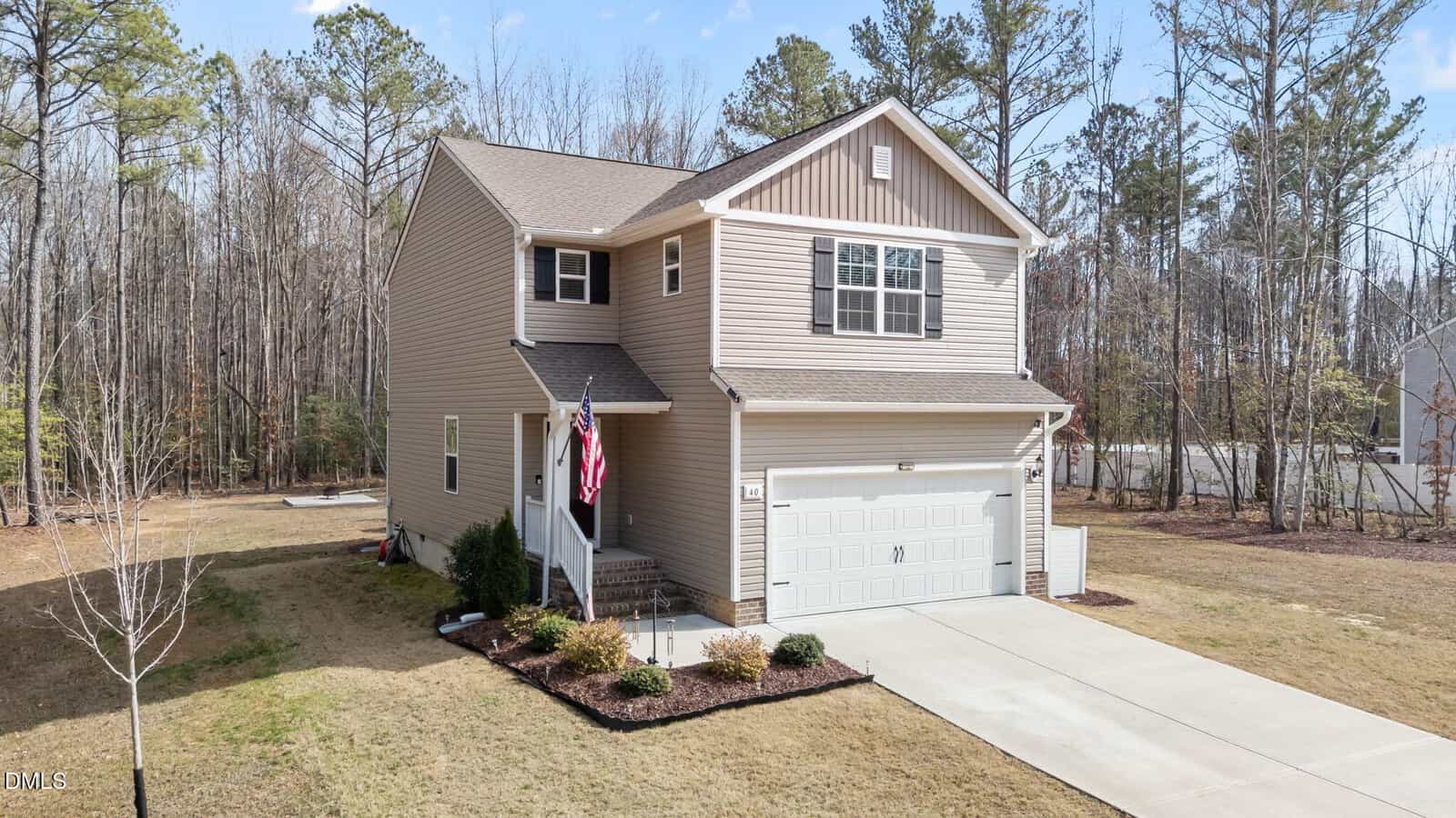 A two-story beige house with a gable roof, white trim, and a double garage. An American flag hangs by the front steps. The yard is partially landscaped, with grass and small shrubs, and is bordered by trees.