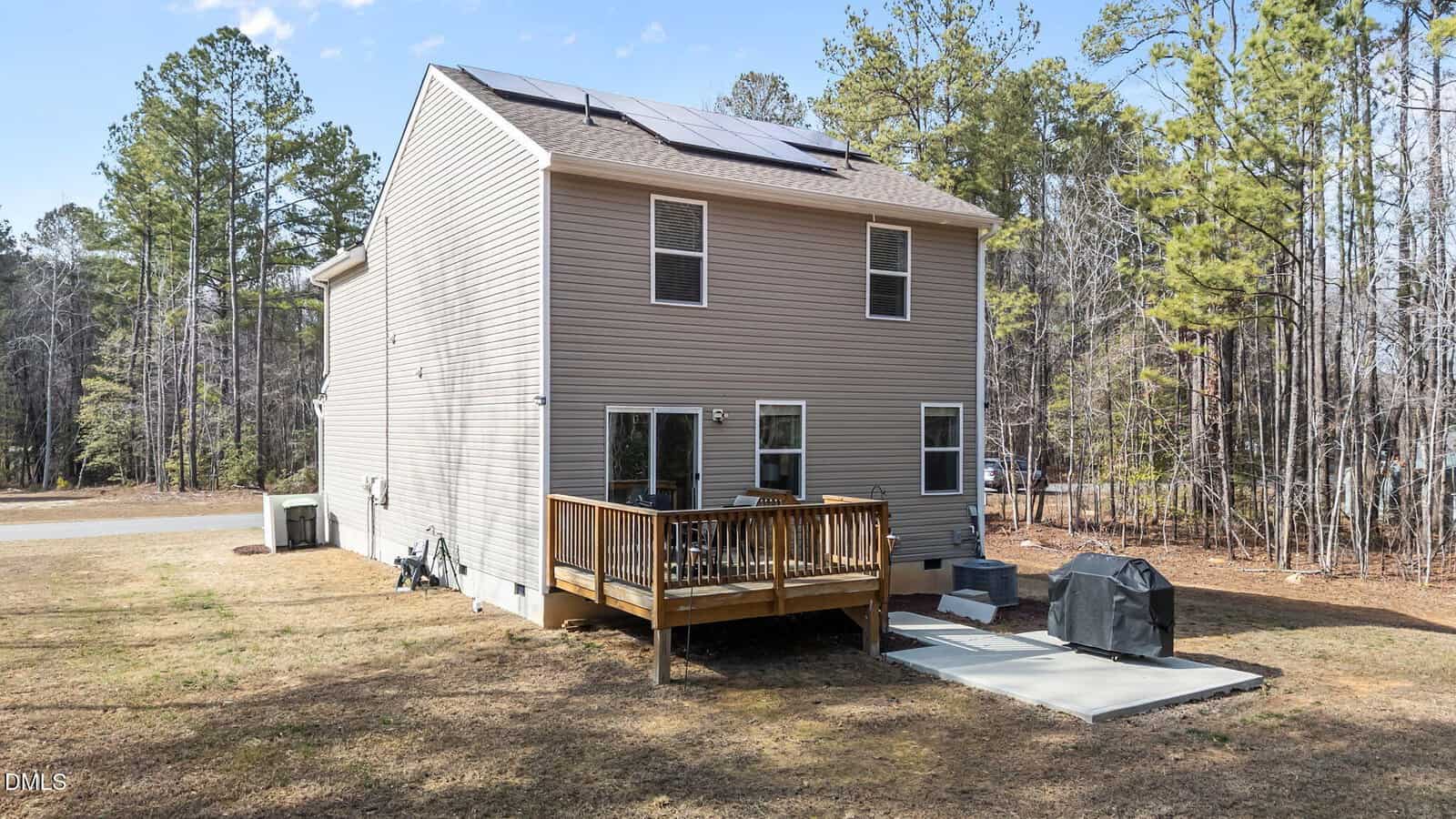 Two-story beige house with solar panels on the roof, a small wooden deck with seating, and a concrete patio with a grill. The backyard is grassy and surrounded by tall trees.