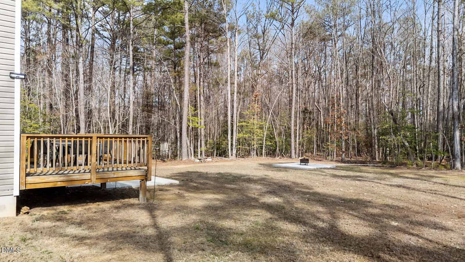 A wooden deck is attached to a house on the left, overlooking a grassy backyard with a small fire pit area. The yard is bordered by a dense forest of tall trees without leaves.