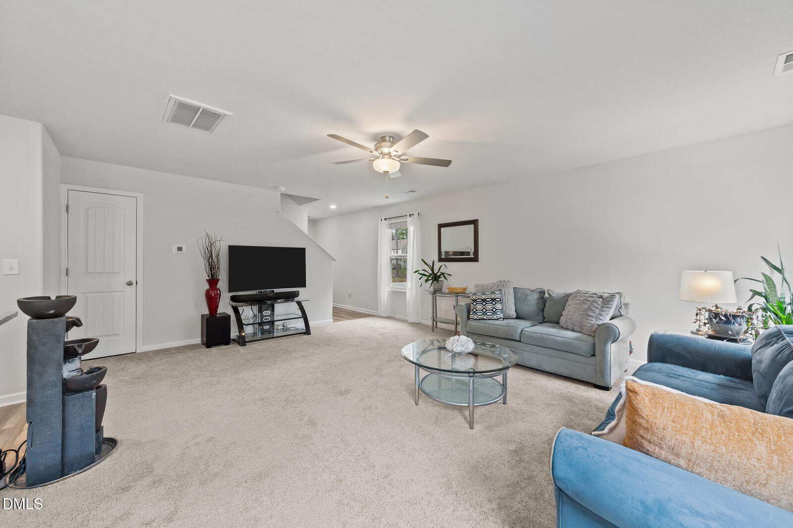 A spacious living room with beige carpet, two blue sofas, a glass coffee table, a flat-screen TV on a stand, indoor plants, and a ceiling fan with lights. A staircase and a door are visible in the background.
