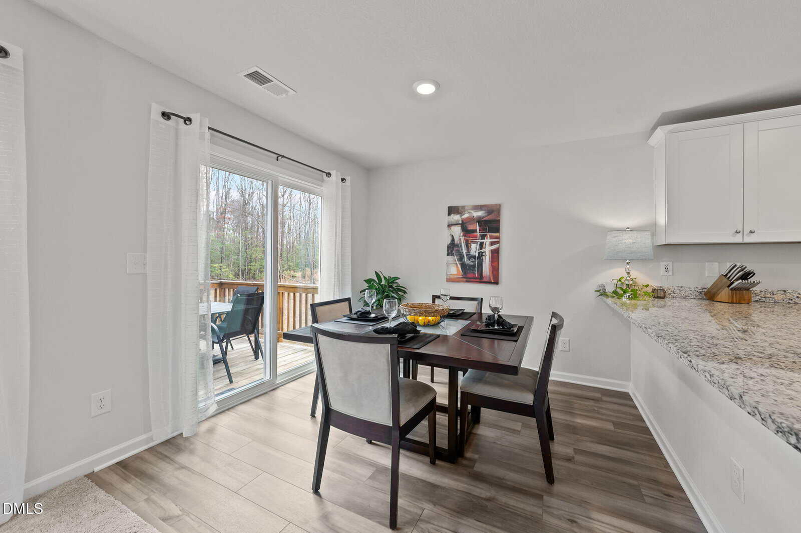 Dining area with a dark wood table and four chairs next to sliding glass doors leading to a deck; modern decor includes a wall art piece, granite countertop, and light wood flooring.