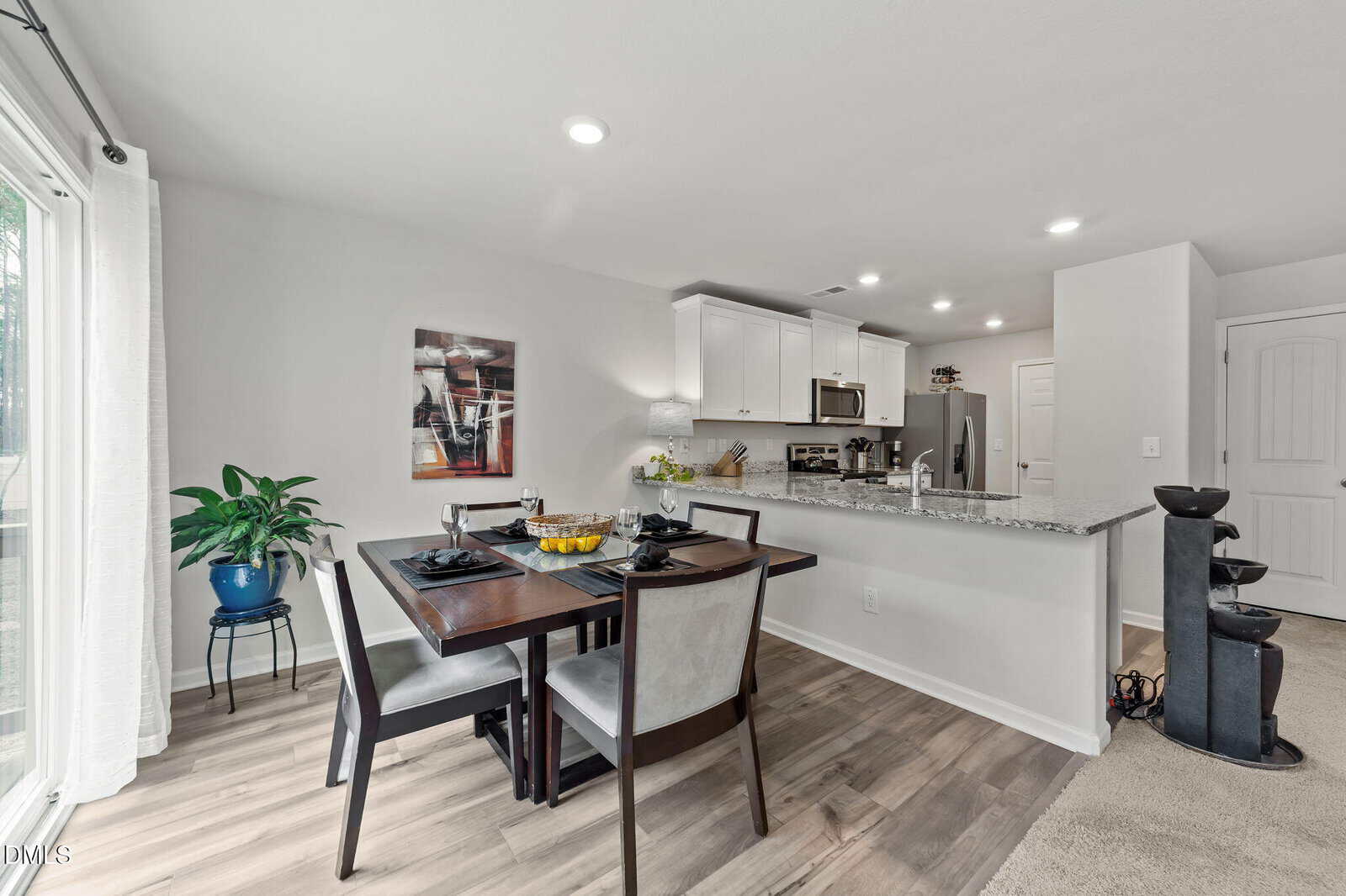 A modern dining area with a dark wood table set for four, next to a kitchen with stainless steel appliances and white cabinets. Light wood flooring, a potted plant, and wall art are visible. Bright, natural light enters from the left.