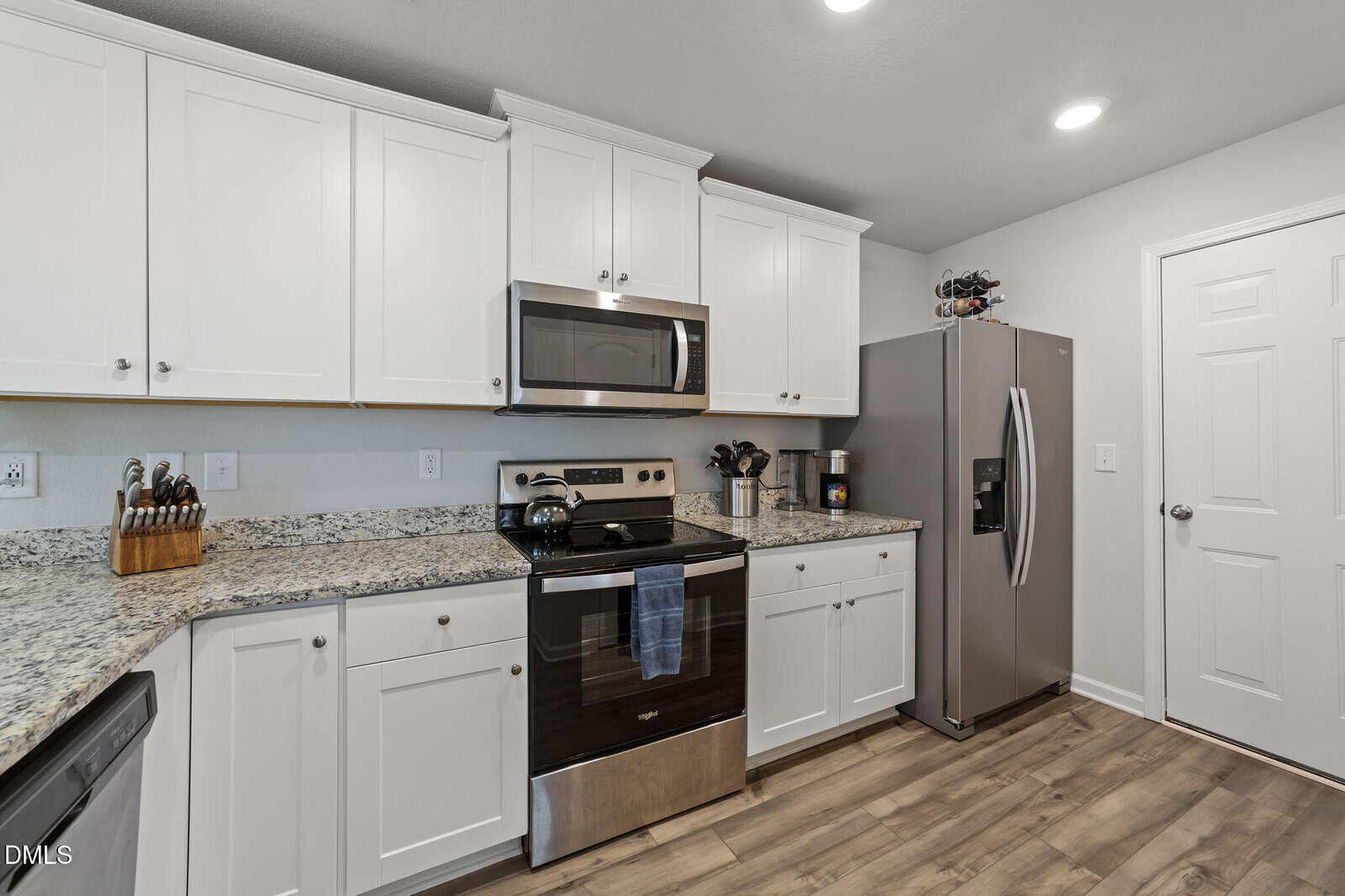 A modern kitchen with white cabinets, granite countertops, stainless steel appliances including a stove, microwave, and refrigerator, and light wood flooring. A knife block and utensils are on the counter.