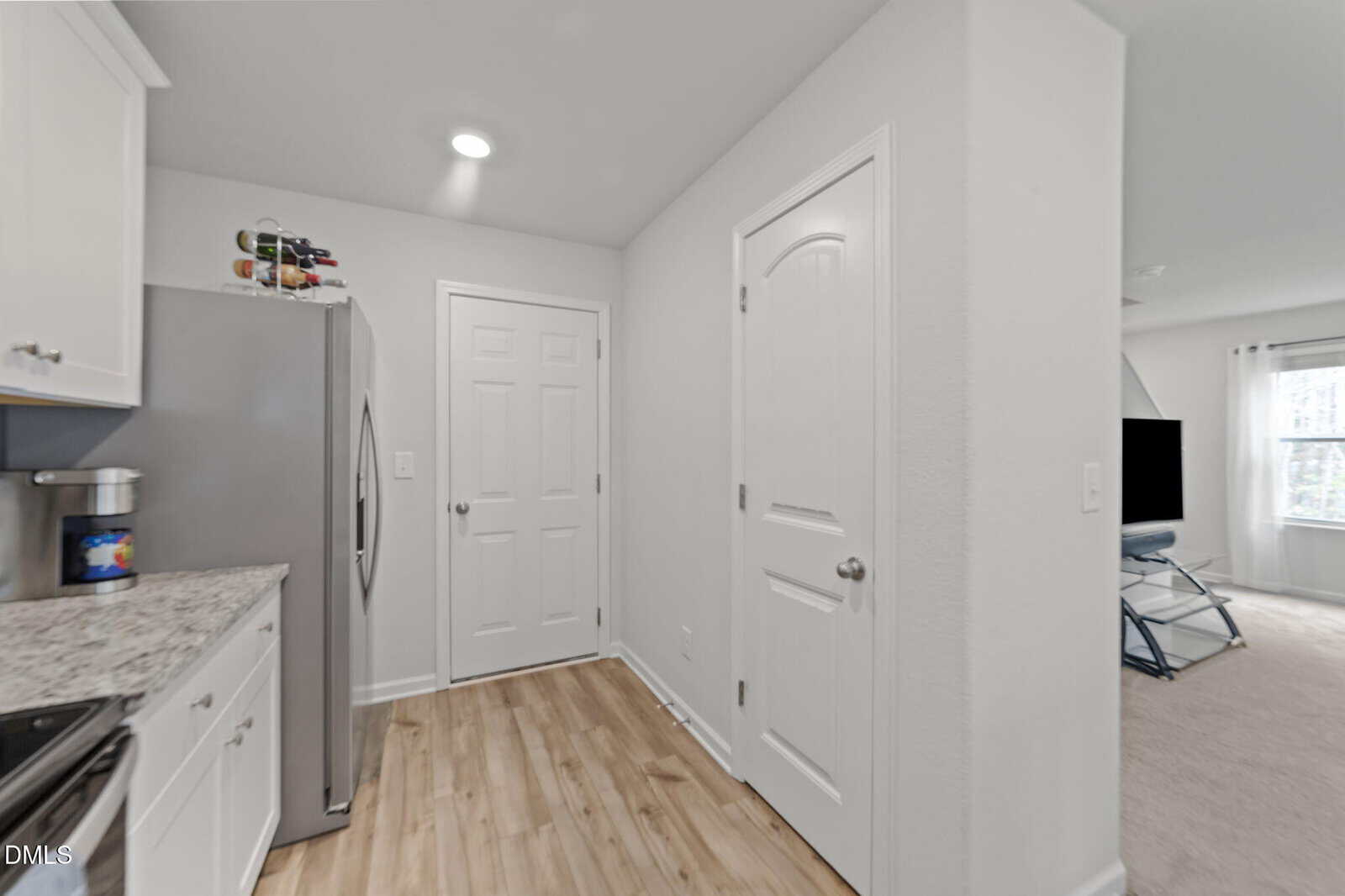 A compact kitchen with white cabinets, stainless steel refrigerator, and light wood floor. Two closed white doors are on the right, and a living area with a TV and window is partially visible in the background.