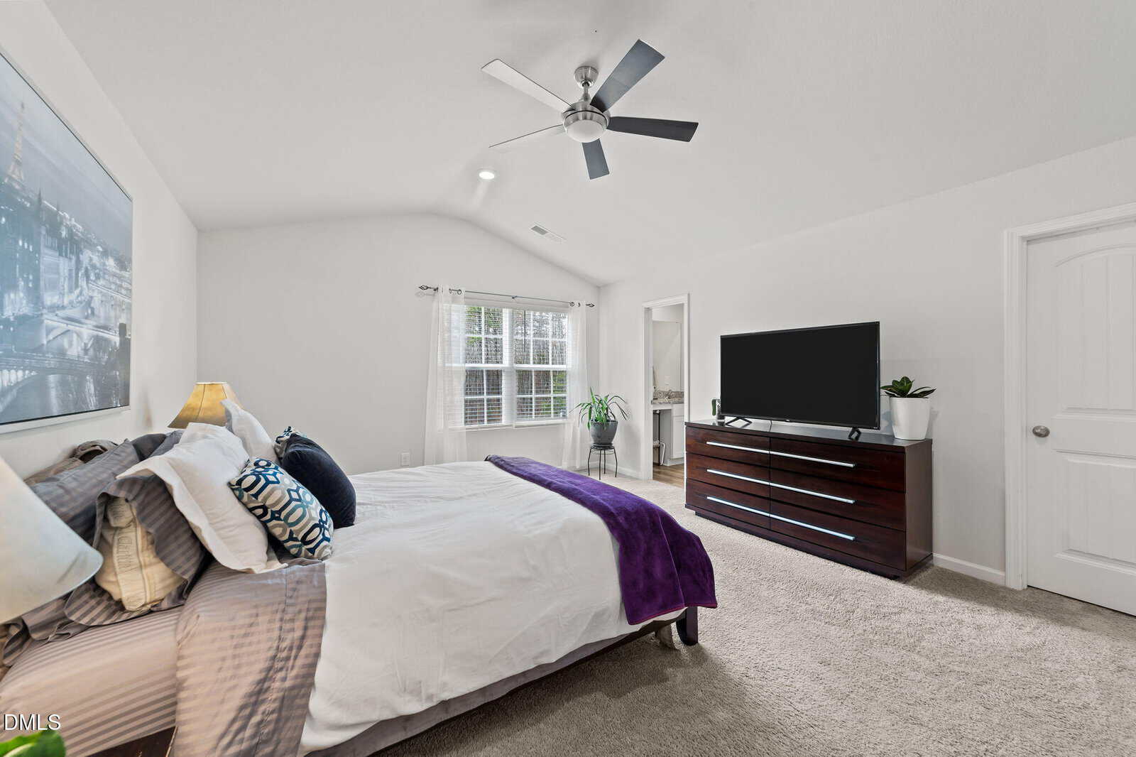 A bedroom with a bed, pillows, and blanket on the left, a large dresser with a flat-screen TV on the right, a ceiling fan above, a window with white curtains, and an open doorway leading to a bathroom.