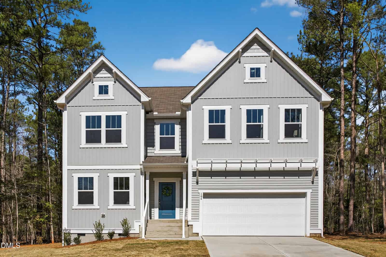 A two-story, light gray house with white trim, multiple windows, a blue front door, and a double garage, set against a backdrop of tall trees under a blue sky.