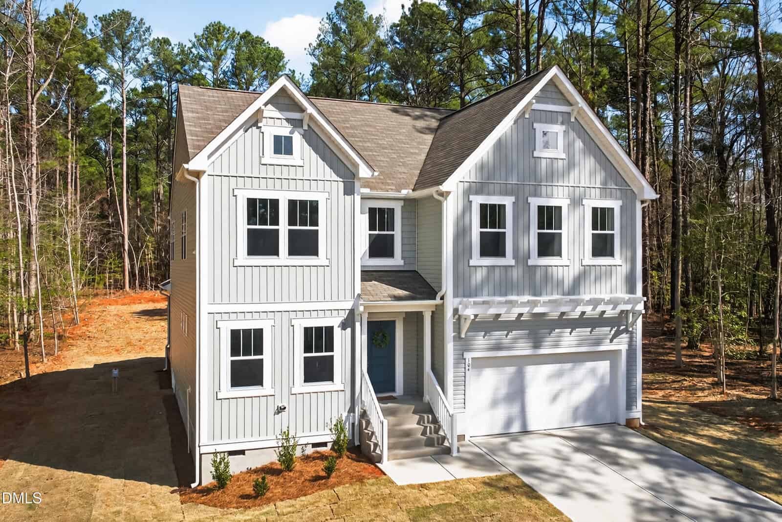 A two-story light gray house with white trim, a covered front porch, and a double garage, set on a grassy lawn with small shrubs and surrounded by tall trees.