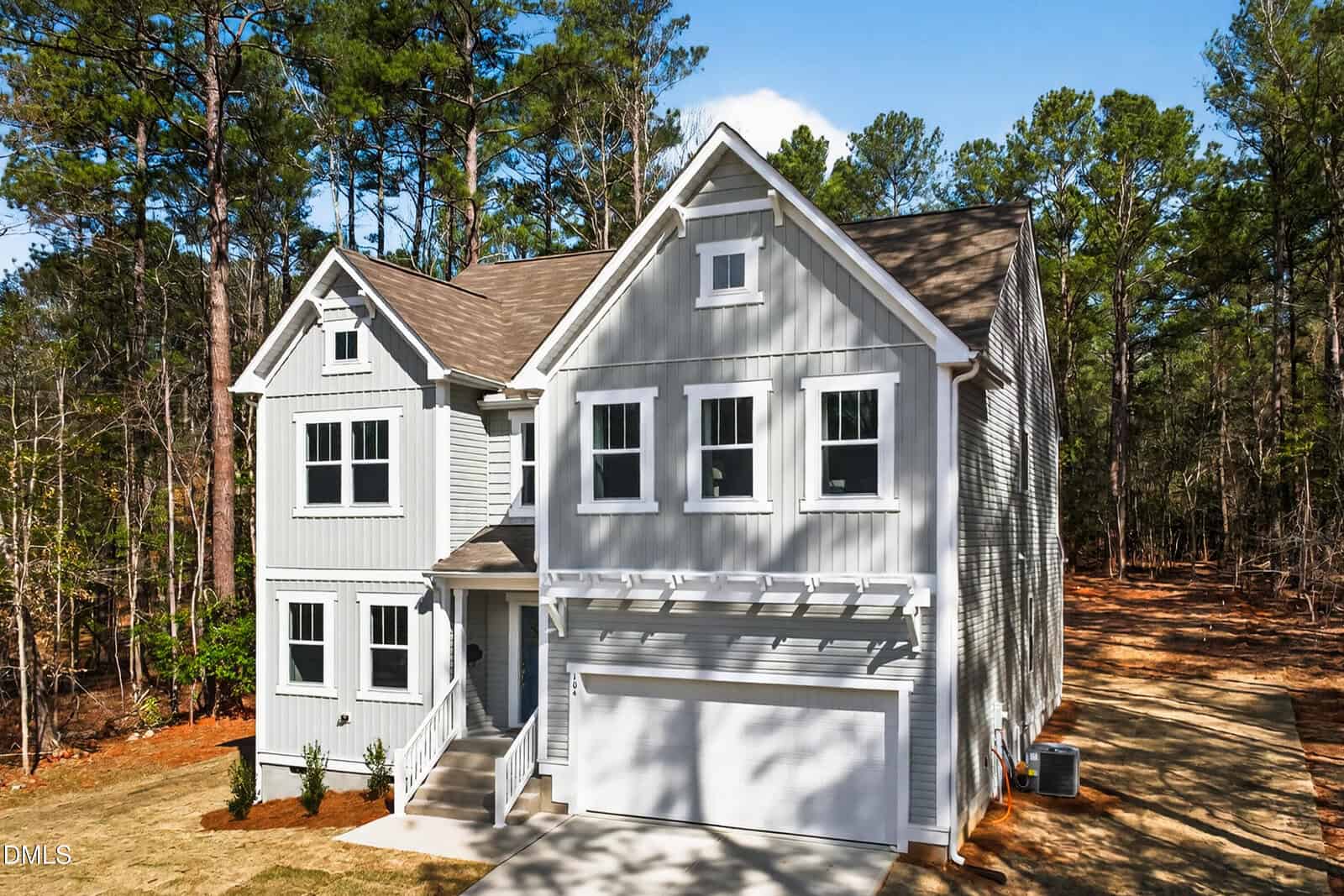 A two-story, light gray house with white trim and a double garage, set on a lot surrounded by tall trees. The driveway and small front yard are visible, and the house sits near a wooded area.