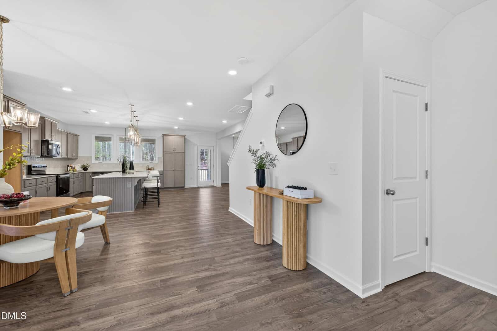 Open-concept kitchen and dining area with wood flooring, round dining table with four chairs, modern kitchen with white cabinets and stainless steel appliances, and a wooden console table near a white door.