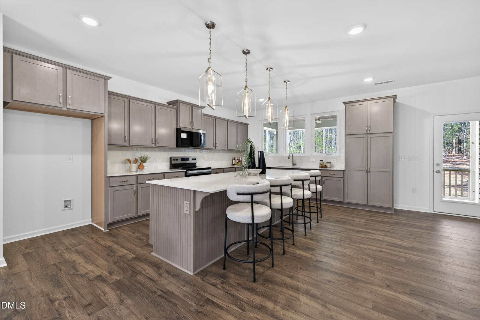 Spacious modern kitchen with light gray cabinets, stainless steel appliances, large island with seating for five, pendant lighting, and wood flooring. Large windows and a glass door provide natural light and outdoor views.