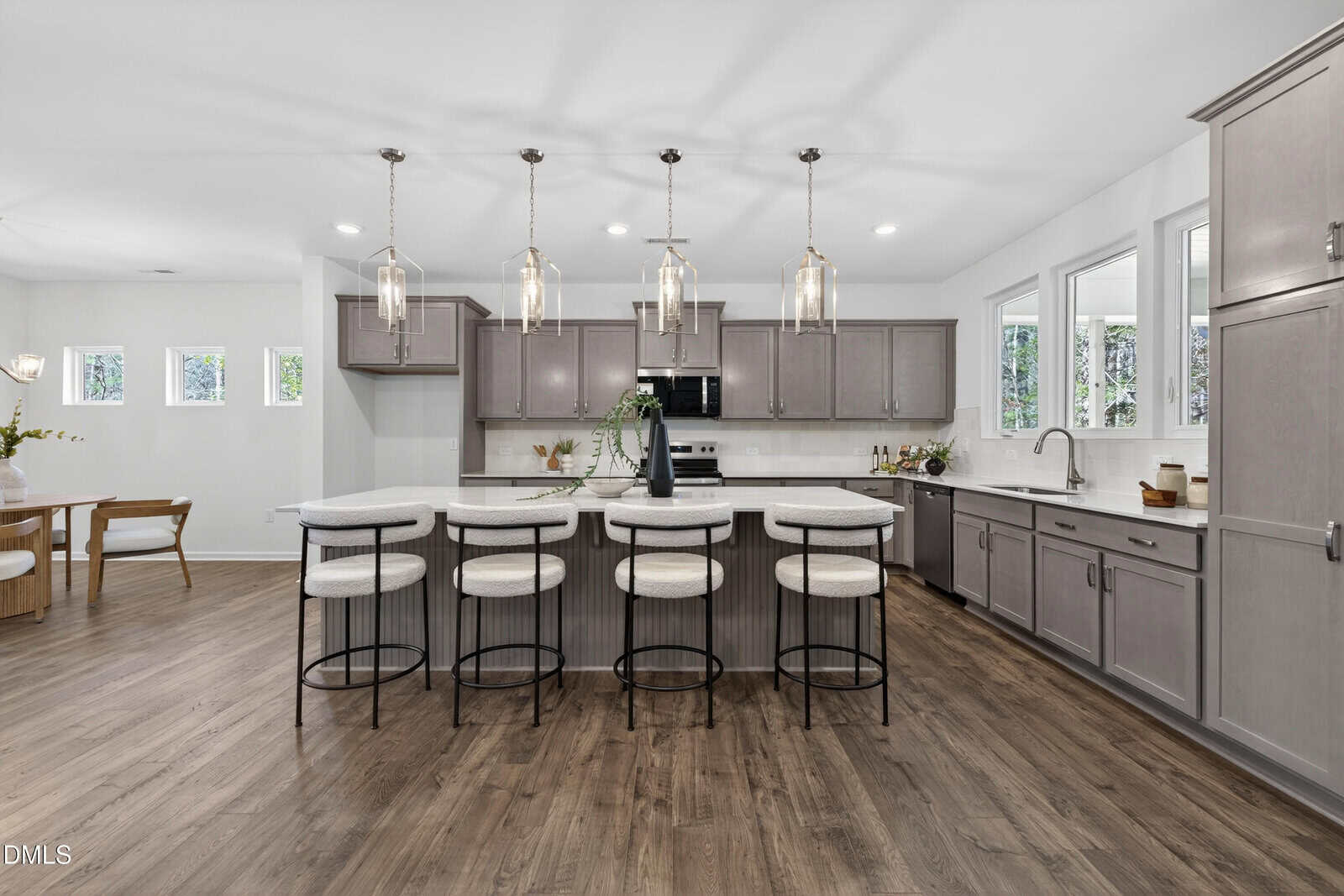 Modern kitchen with gray cabinets, white countertops, four cushioned bar stools at an island, pendant lights above, stainless steel appliances, and wood flooring. Dining area visible in the background.