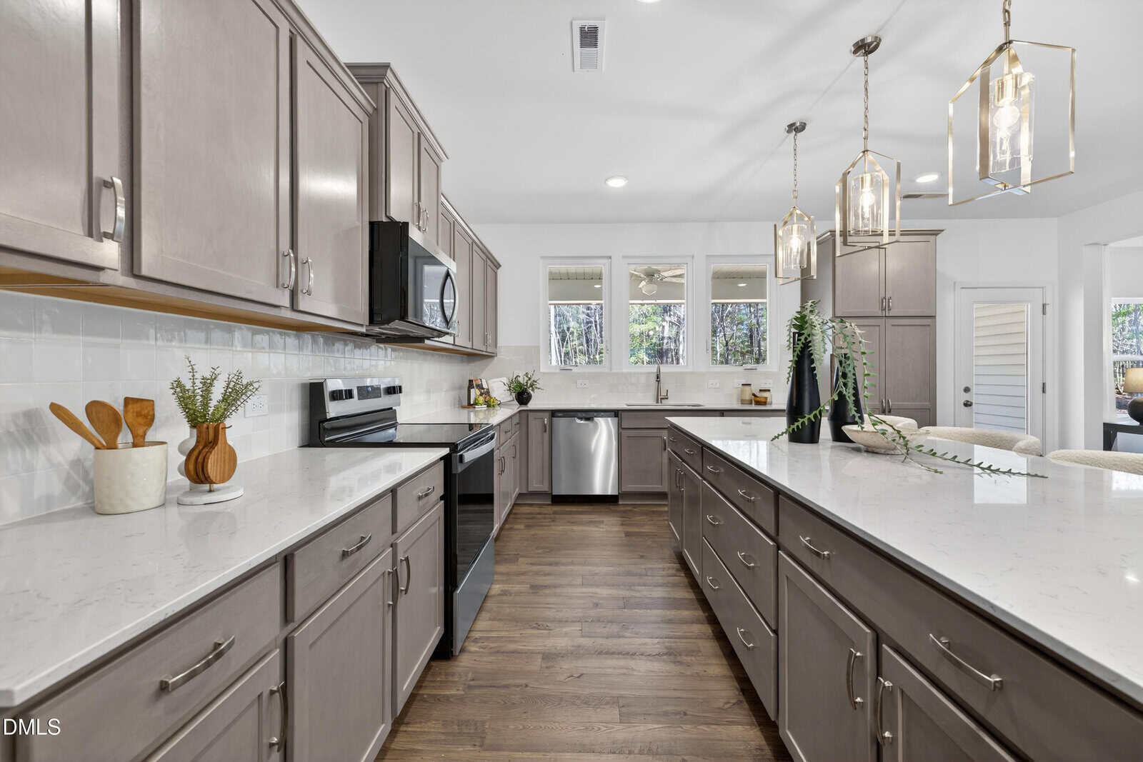 Modern kitchen with light gray cabinets, white countertops, stainless steel appliances, and pendant lights over a large island. Hardwood floors, decorative items, and windows provide a bright, clean atmosphere.