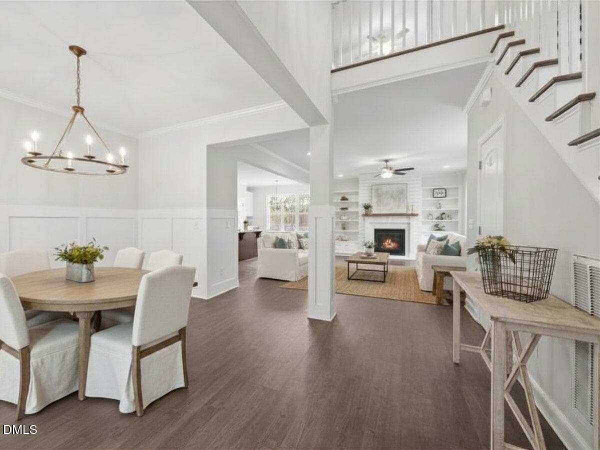 Open-concept living and dining area with white walls, wood flooring, round dining table with four chairs, cozy living room with fireplace, and a staircase leading upstairs. Natural light fills the space.