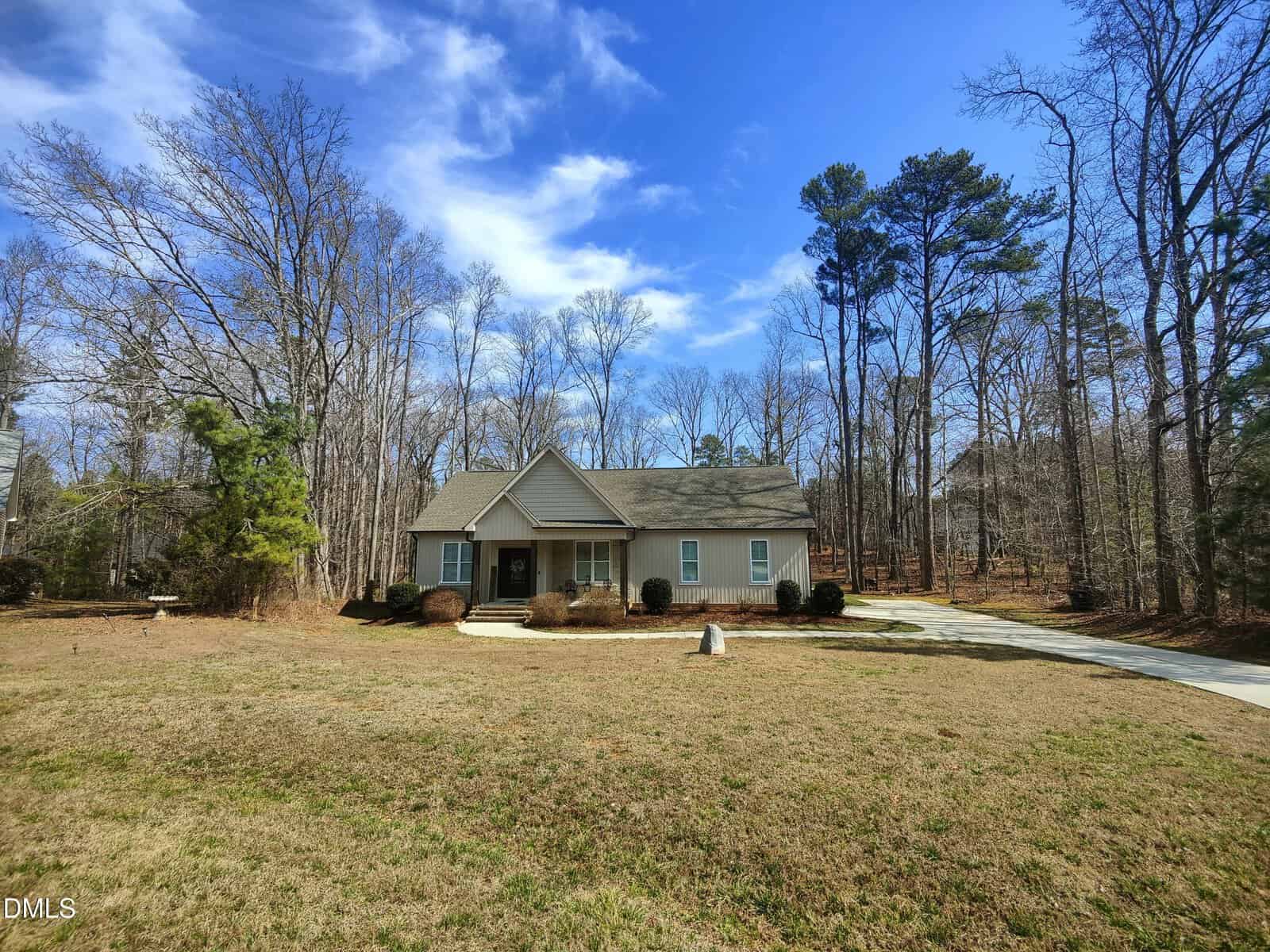 Single-story beige house with a gabled roof, front porch, and attached garage sits on a large grassy lot surrounded by tall, leafless trees under a blue sky with scattered clouds. A curved driveway leads up to the house.