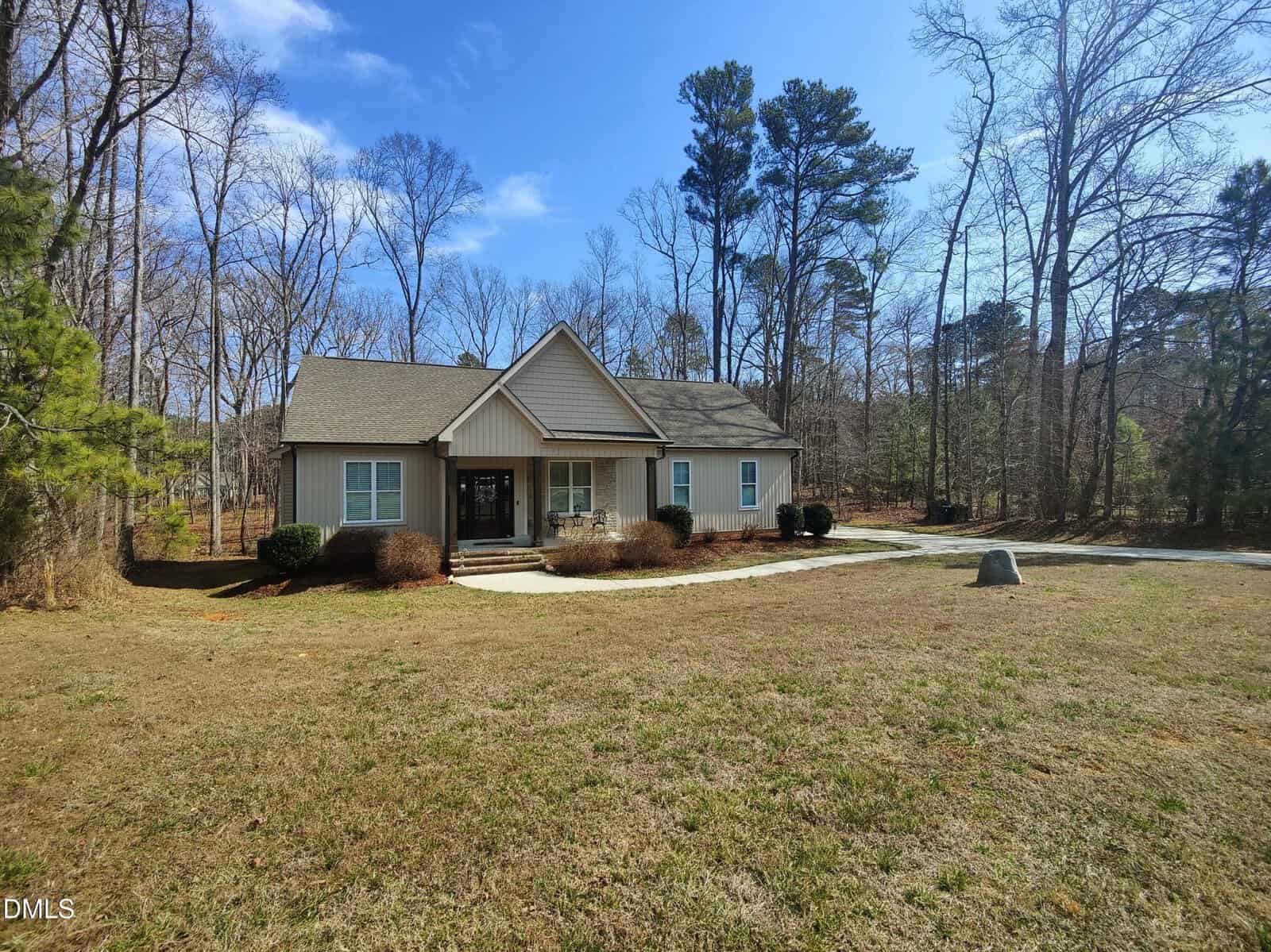 A single-story beige house with a gable roof sits in a large, grassy yard bordered by trees under a blue sky. A concrete walkway leads to the front door, and a driveway is visible on the right.