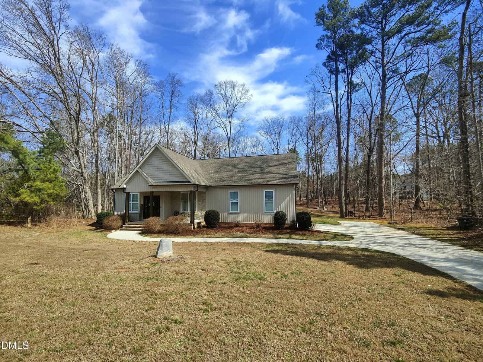 A single-story house with beige siding and a covered front porch sits in a clearing surrounded by trees, with a curved driveway and a grassy yard under a partly cloudy sky.