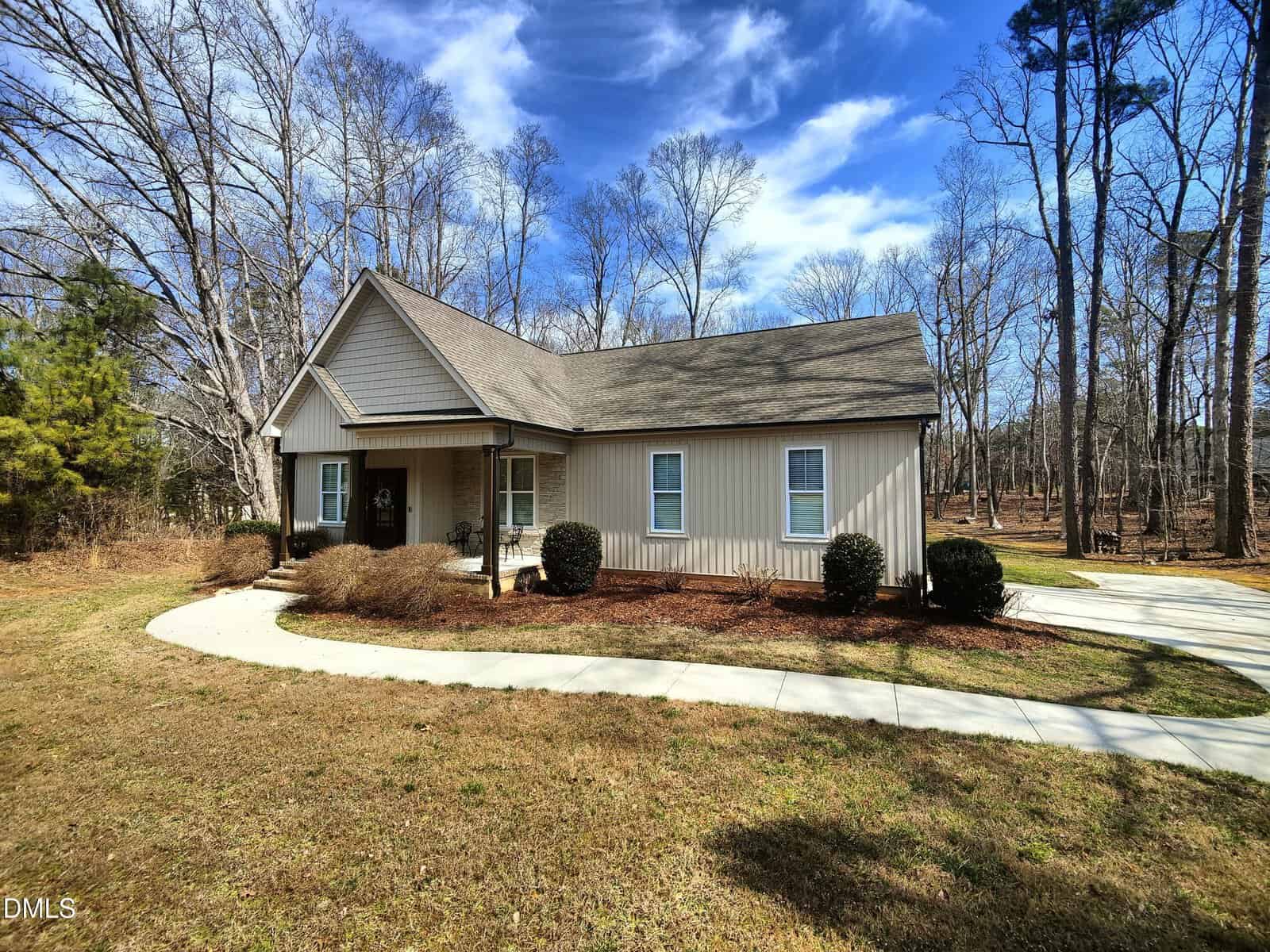 A single-story beige house with a gable roof and a covered front porch, surrounded by leafless trees and landscaped shrubs, features a curved driveway and a lawn under a partly cloudy blue sky.