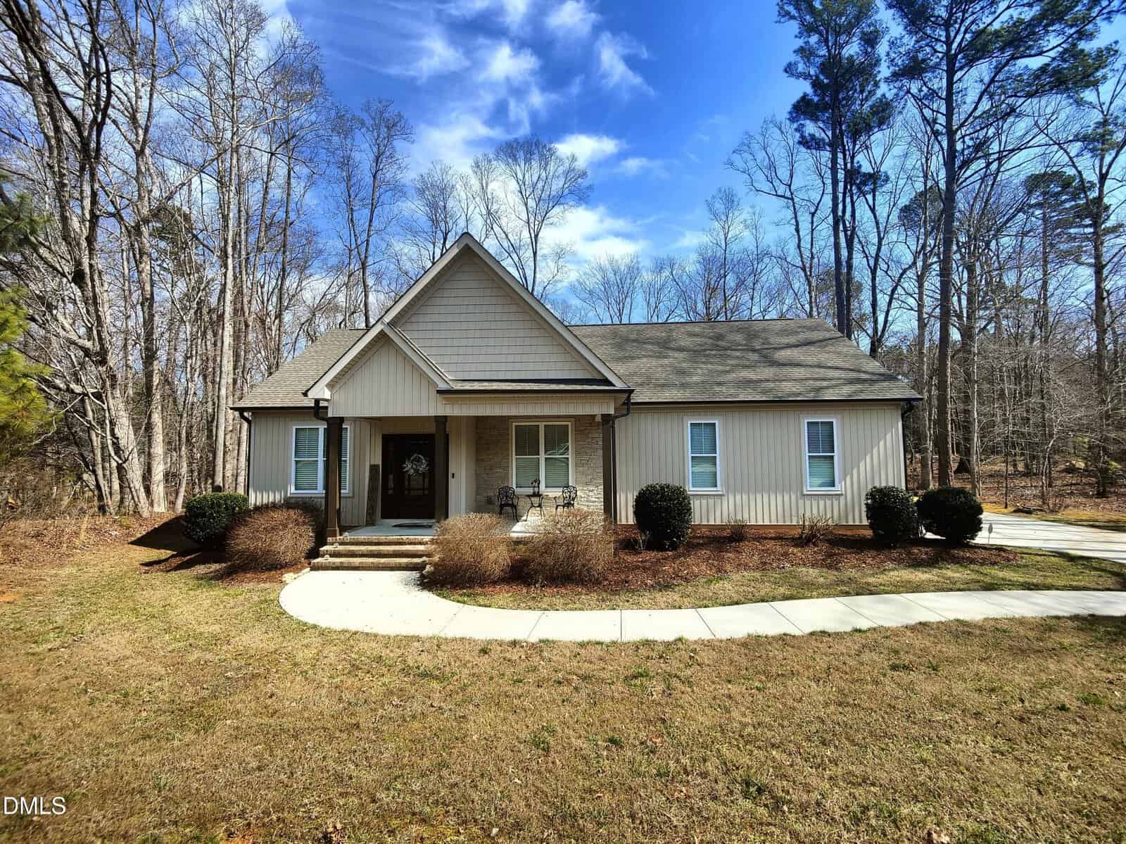 A single-story beige house with a gabled roof, front porch, and white trim sits amid a grassy yard with shrubs and a curved driveway, surrounded by tall trees under a blue sky with scattered clouds.