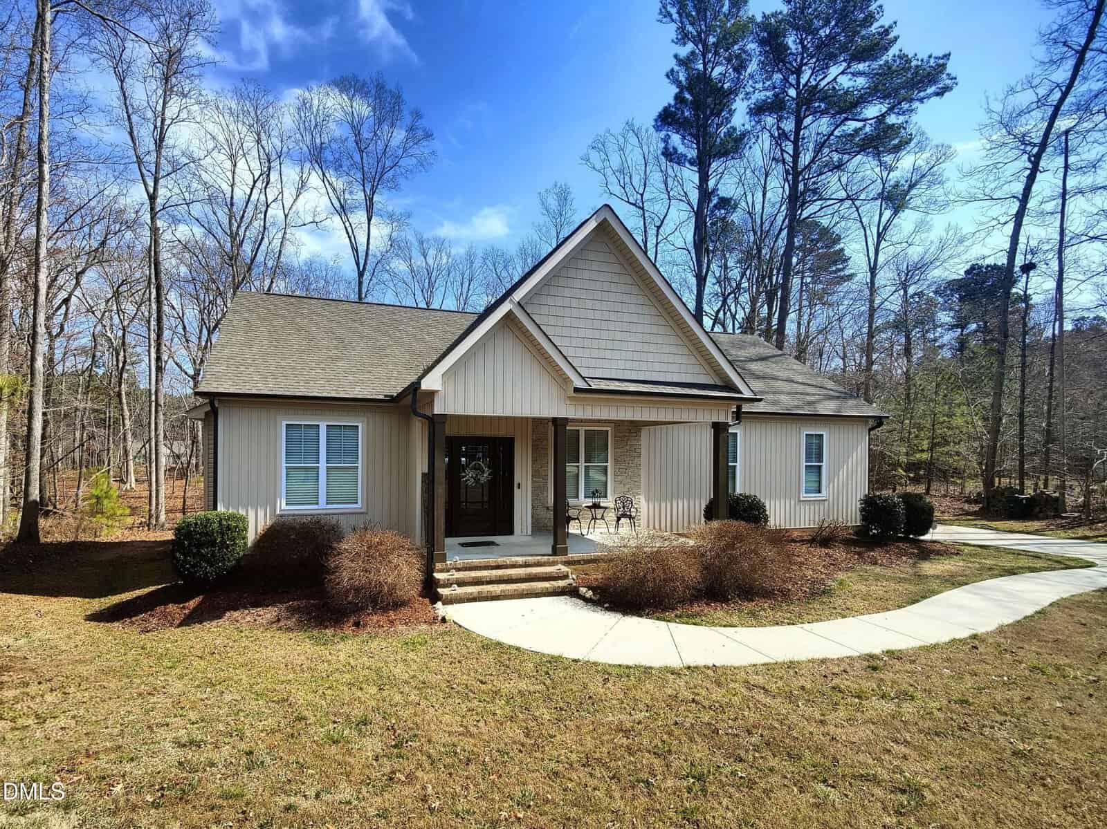 Single-story house with beige vertical siding, gable roof, covered front porch, and two front-facing windows. A curved concrete walkway leads to the entrance. Leafless trees and shrubs are in the yard. Blue sky with some clouds.