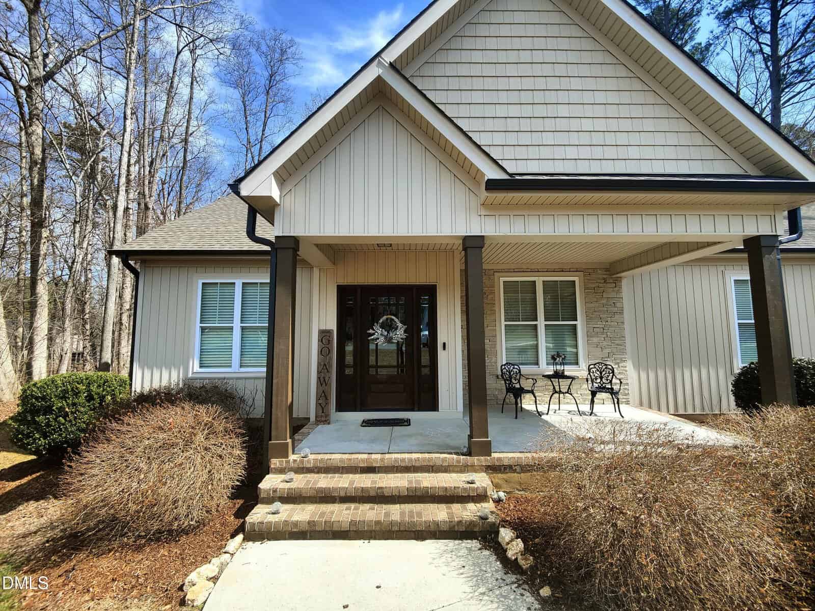 Single-story beige house with a covered front porch, dark brown columns, a glass front door, and two black metal chairs with a small table. Leafless trees and shrubs surround the entryway. Steps lead up to the porch.