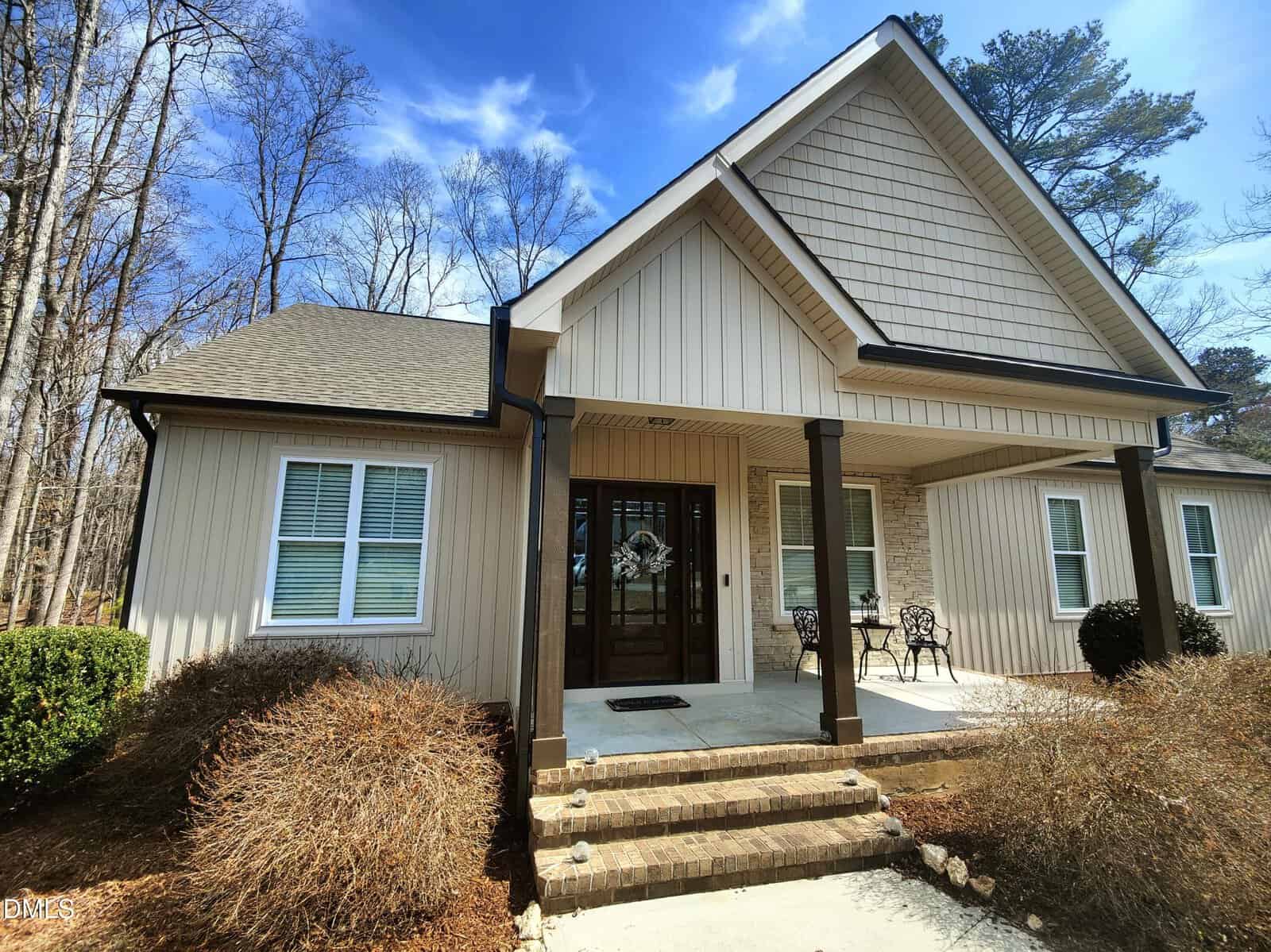 Single-story beige house with vertical siding, dark trim, two front windows with shutters, and a covered entryway supported by columns. There are two chairs near the front door and leafless shrubbery along the walkway.