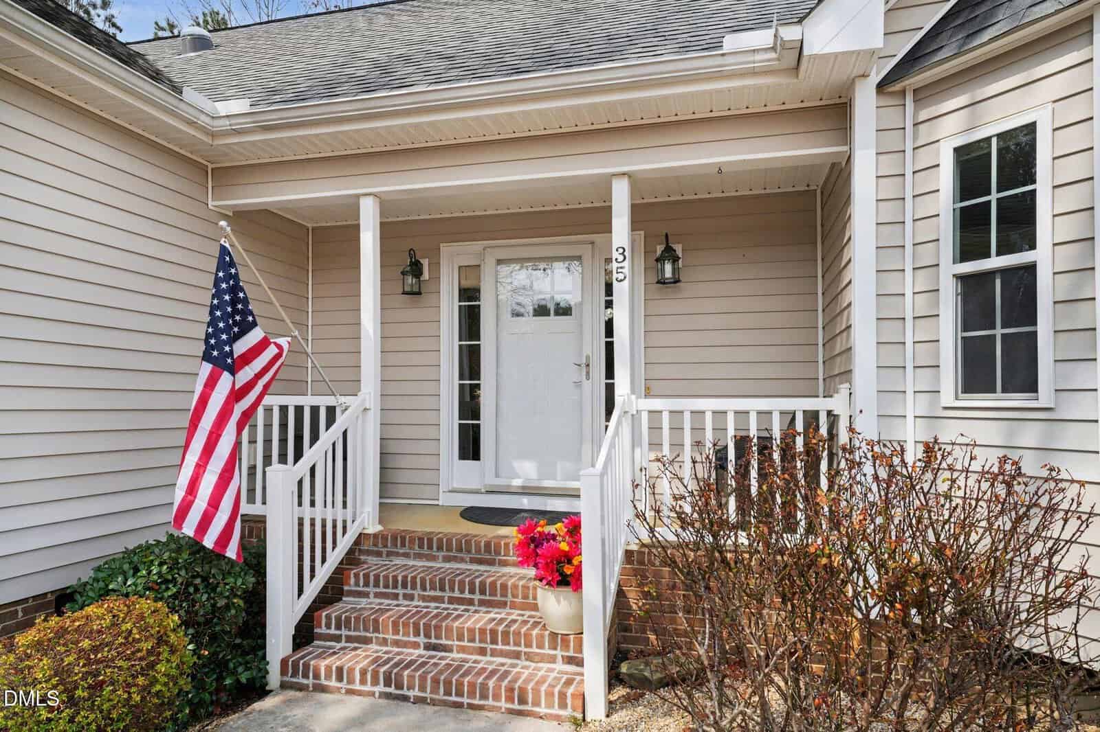 A front porch with brick steps, white railing, a glass door, wall-mounted lights, a house number 35, an American flag, a potted plant with red flowers, and shrubs around the entrance.