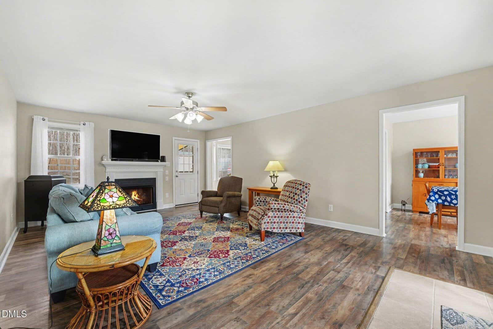 A living room with a blue sofa, patterned chair, area rug, and wicker side table. A ceiling fan is overhead, a TV is above a fireplace, and there is a door and windows letting in natural light.