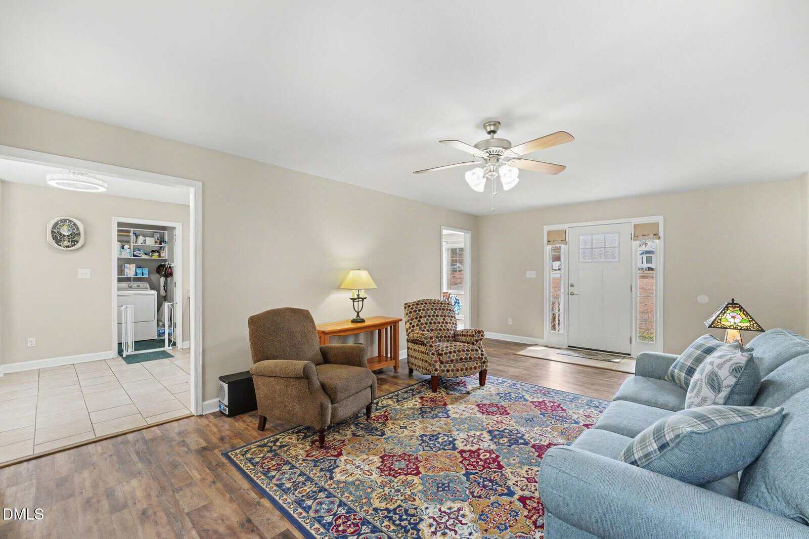 A living room with a patterned rug, blue sofa, two armchairs, wooden side tables with lamps, ceiling fan with lights, and an entry door. The adjacent room has tile flooring and laundry appliances.
