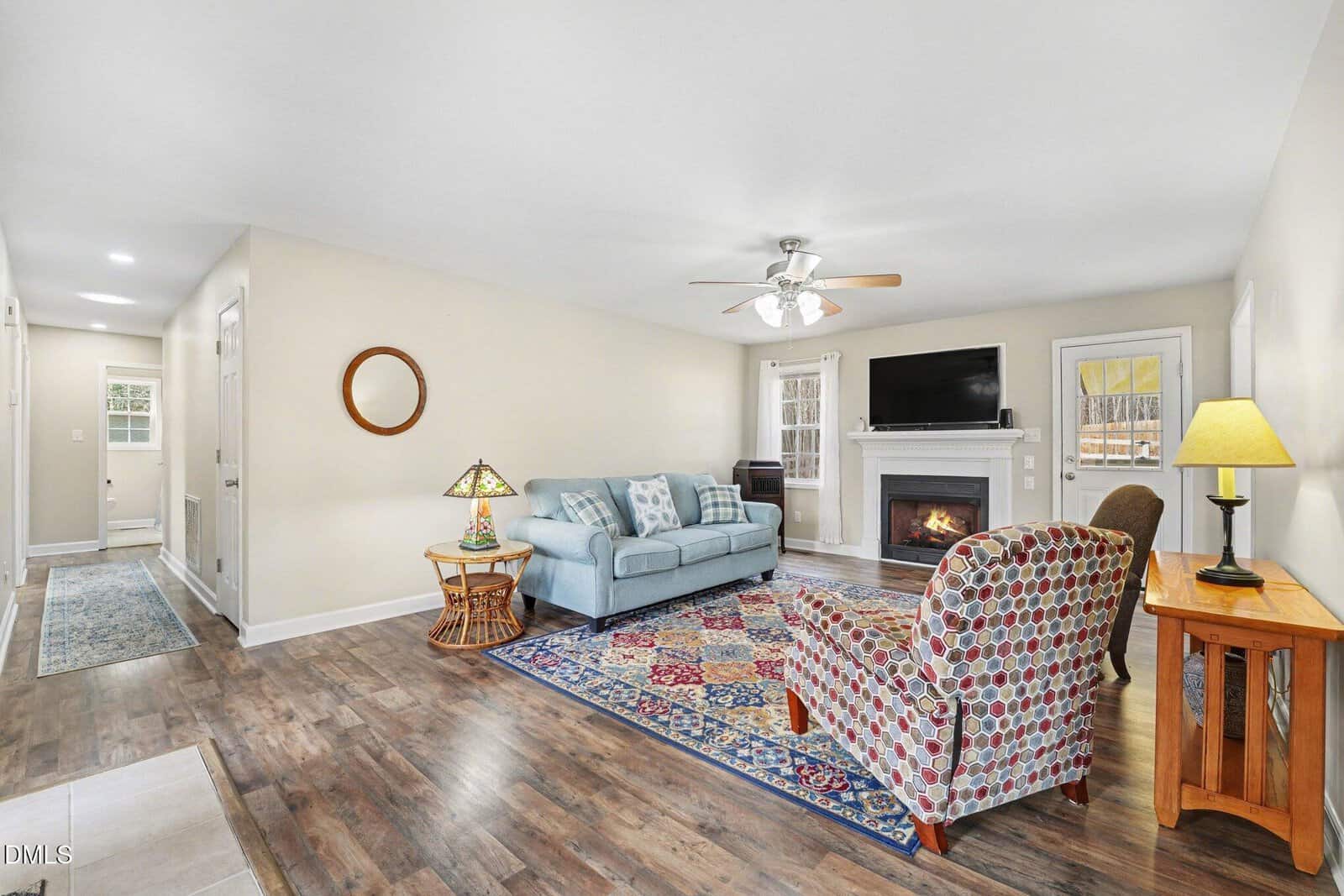 Living room with light walls, wood floor, blue sofa, patterned armchair, area rug, fireplace with TV above, ceiling fan, side tables with lamps, and hallway leading to a door. Window and glass door let in natural light.