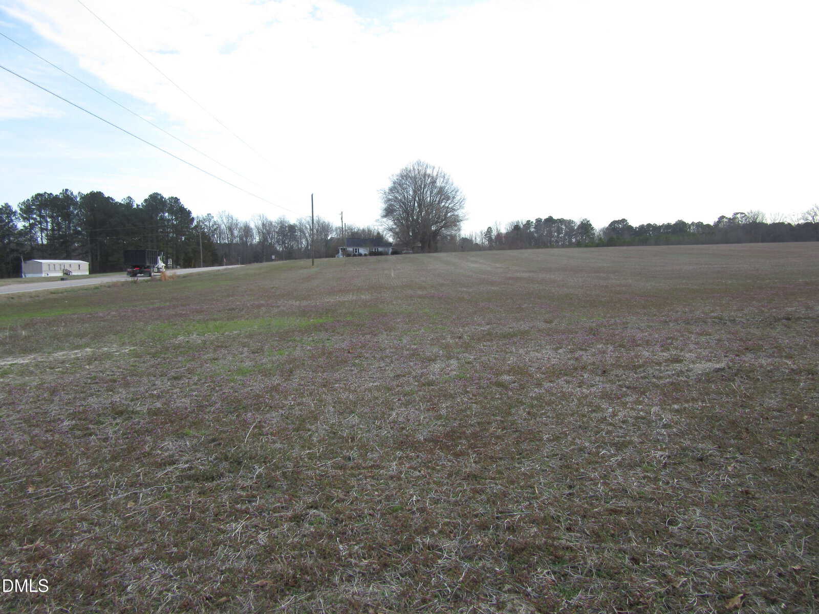 A large, flat, grassy field with sparse vegetation, bordered by trees in the background; a mobile home and a few utility poles are visible near a road on the left side. The sky is mostly cloudy.