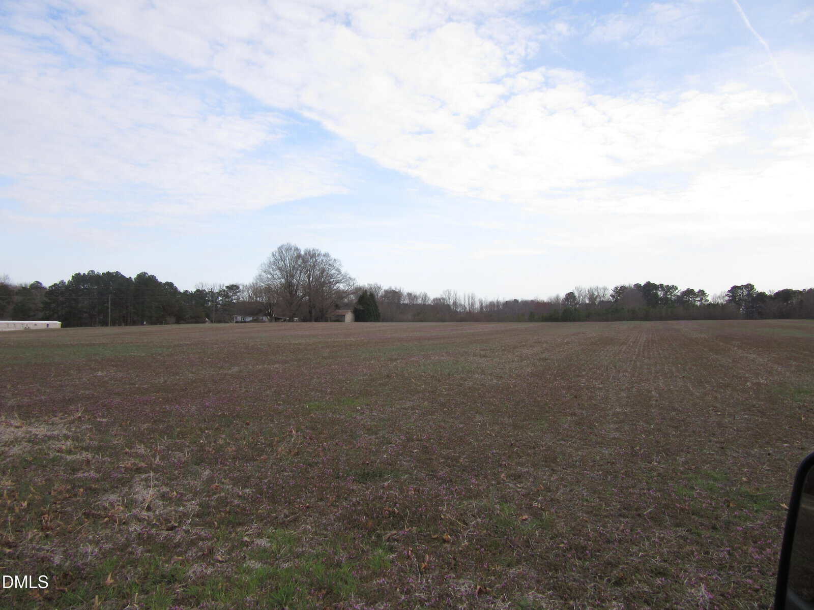 A wide, flat field with short grass and patches of bare soil under a partly cloudy sky. Trees and a few buildings are visible in the distance along the horizon.