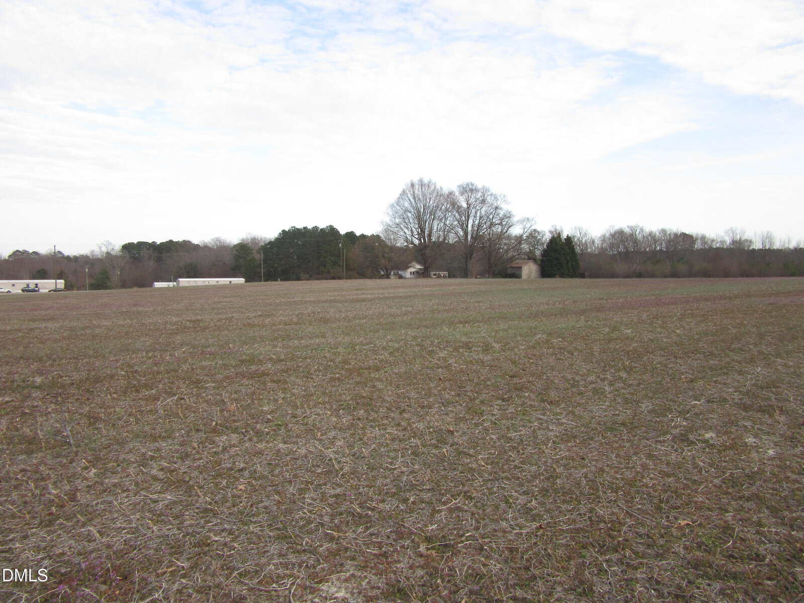A large, open field with sparse grass and patches of bare earth under a mostly cloudy sky. Trees and a few buildings are visible in the distance near the horizon.