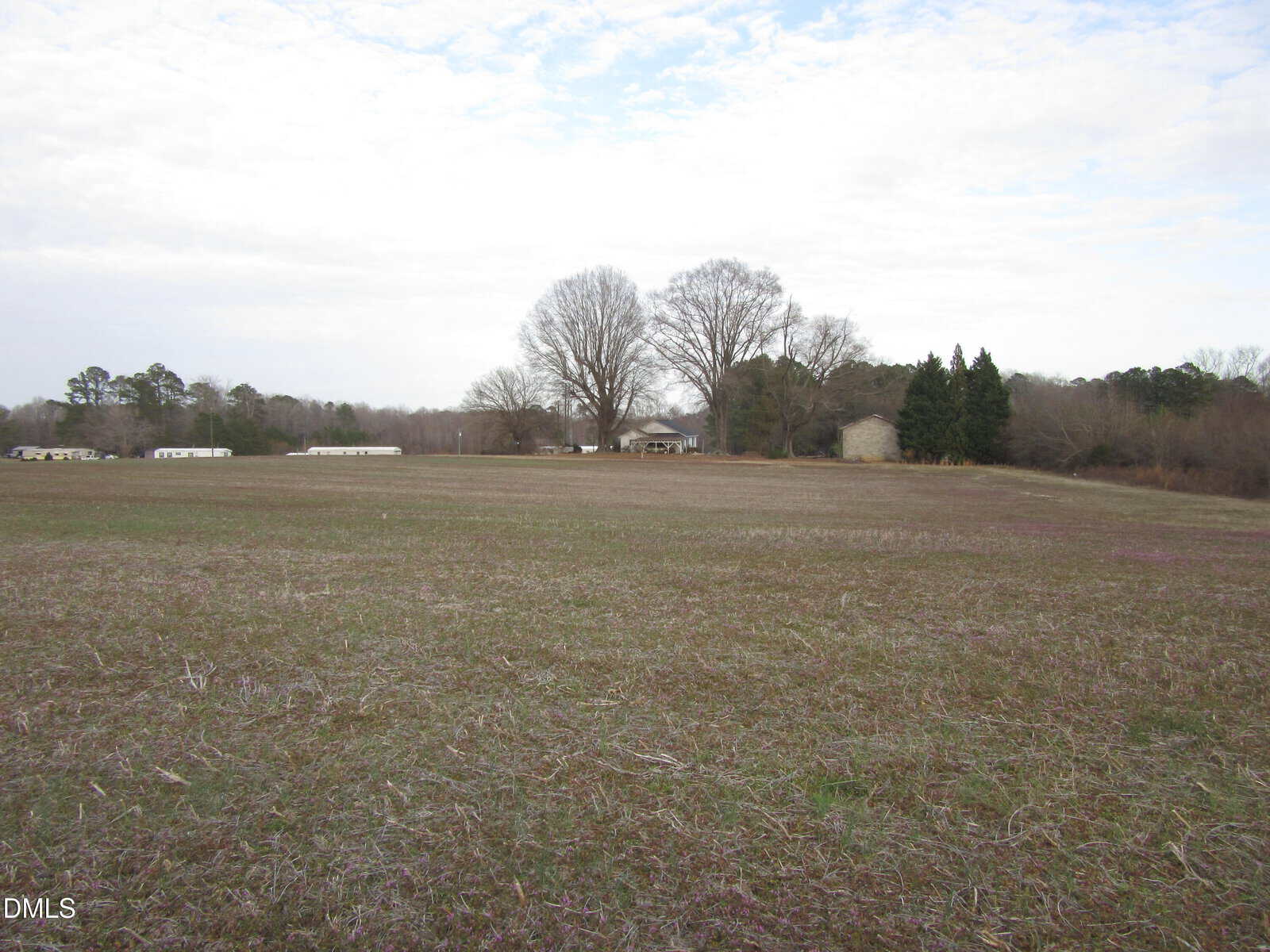 A wide, open field with sparse grass and patches of bare soil, bordered by trees and a few houses in the background under a partly cloudy sky.