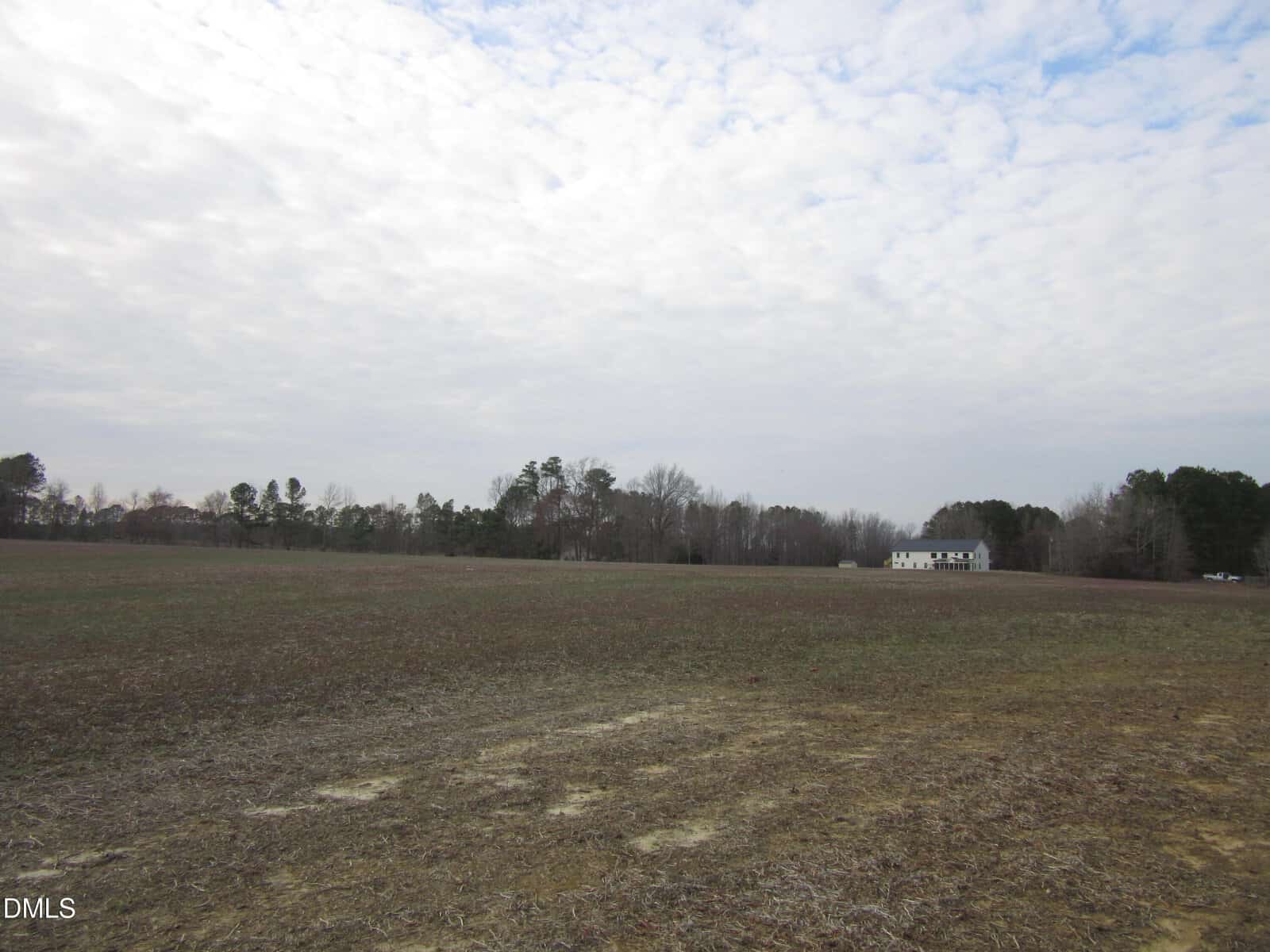 A wide, mostly barren field with patchy grass under a cloudy sky. Trees border the horizon, and a white house is visible in the distance near the right side.