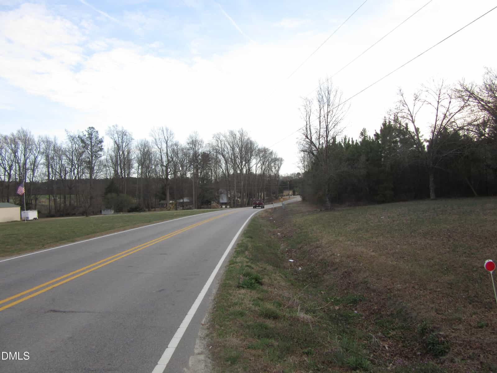 A two-lane rural road curves left, bordered by grass and leafless trees under a partly cloudy sky. Houses and cars are visible in the distance.