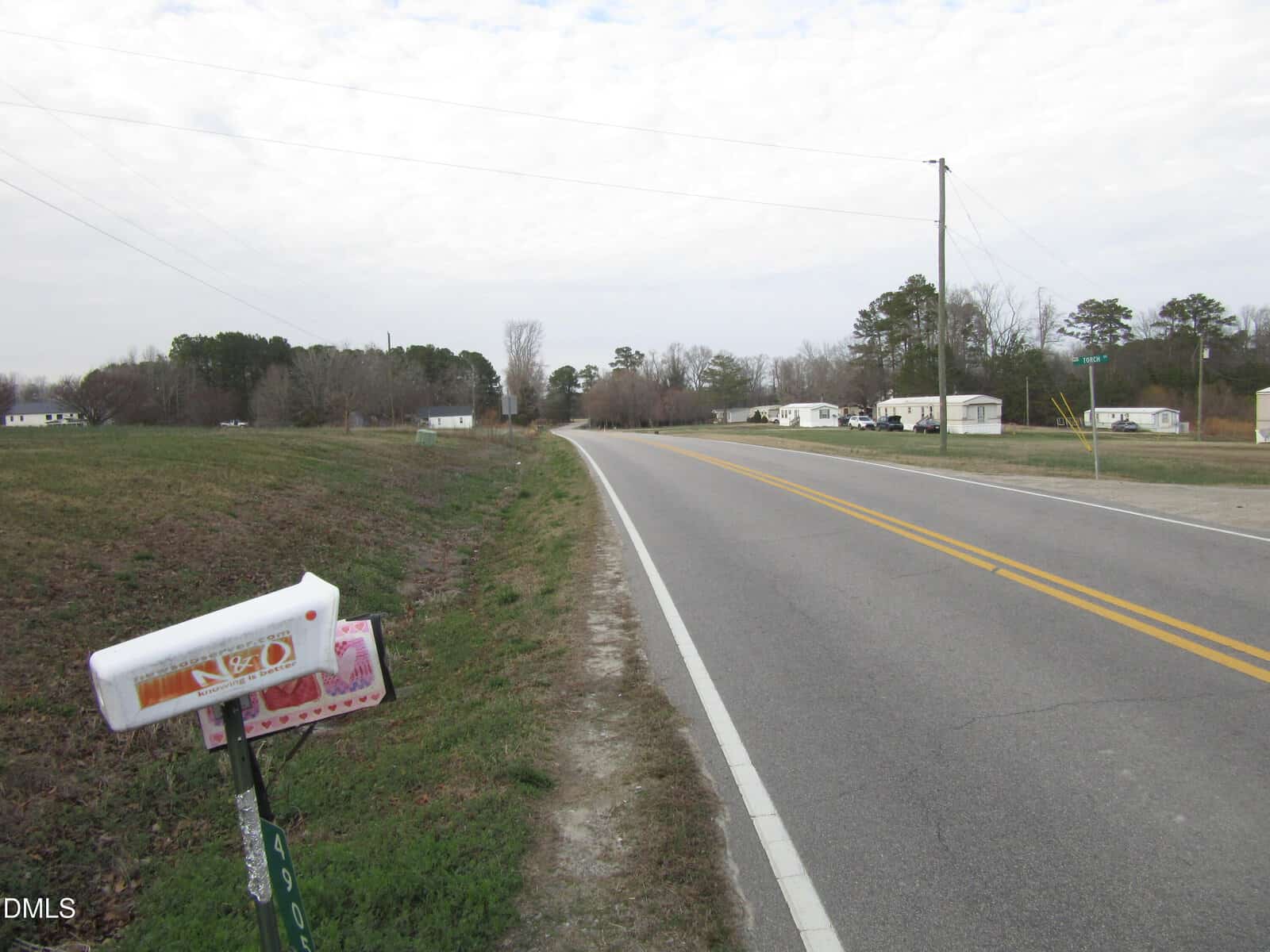 A rural two-lane road curves past fields and scattered trees, with a mailbox in the foreground and mobile homes visible along the roadside under a cloudy sky.
