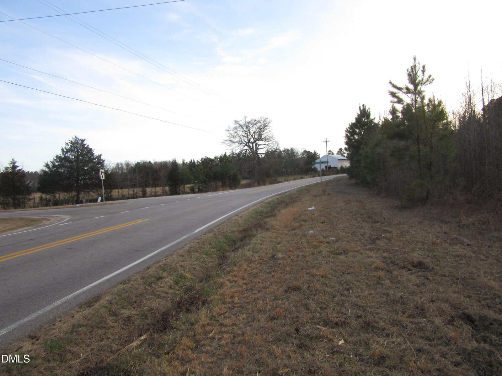 A two-lane rural road curves gently to the left, bordered by grass and trees on both sides under a partly cloudy sky. Power lines run parallel to the road. A white building is visible in the distance.